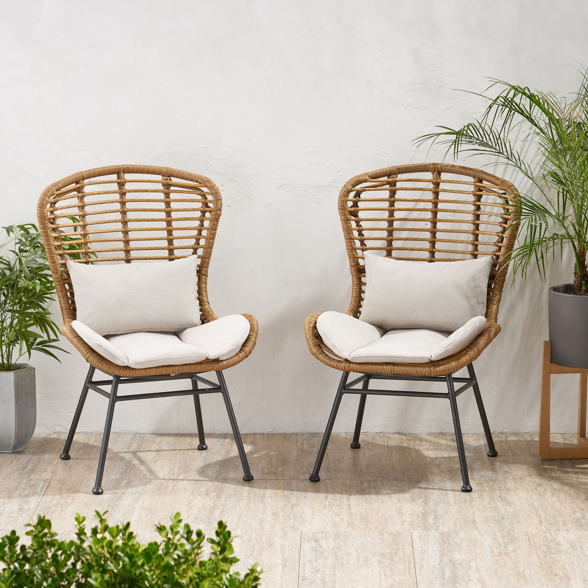 Two Calypso outdoor chairs with water-resistant cushions and iron frames sit side by side on a light tiled floor, surrounded by green potted plants against a white wall.
