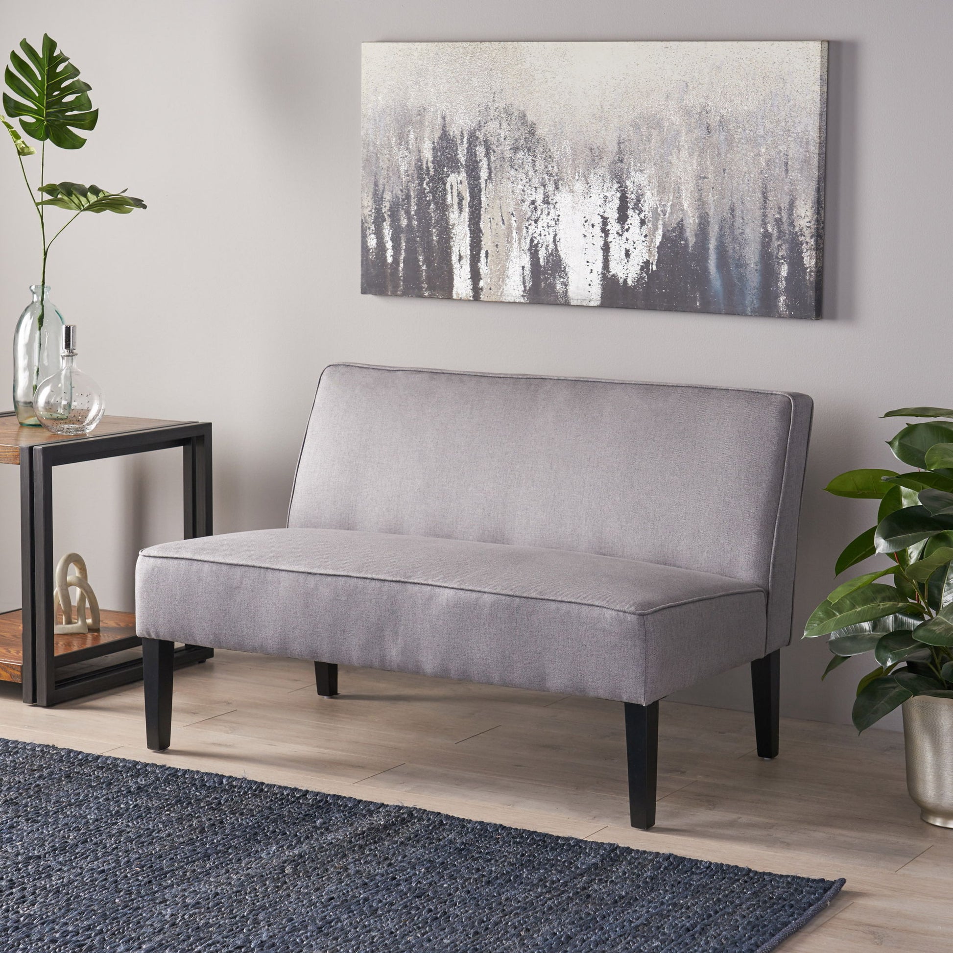 A modern living room featuring a Contemporary Loveseat With Upholstered Fabric And Birch Wood Legs in light gray, a navy textured rug, potted plants, a glass vase, and abstract art on the beige wall above the loveseat.