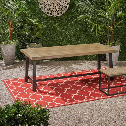 A gray rustic metal outdoor dining table sits on a red patterned rug, surrounded by lush green potted plants, with a decorative wall piece and gravel ground enhancing the garden setting.