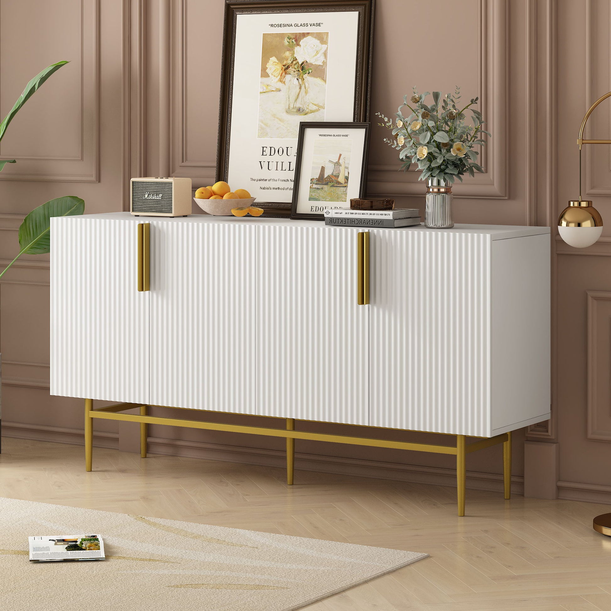 The Modern Elegant 4 Door Sideboard Gold Metal Handle Buffet Cabinet stands against a taupe wall, topped with framed art, a flower vase, oranges in a bowl, and decor items. A beige rug is partly visible on the floor.