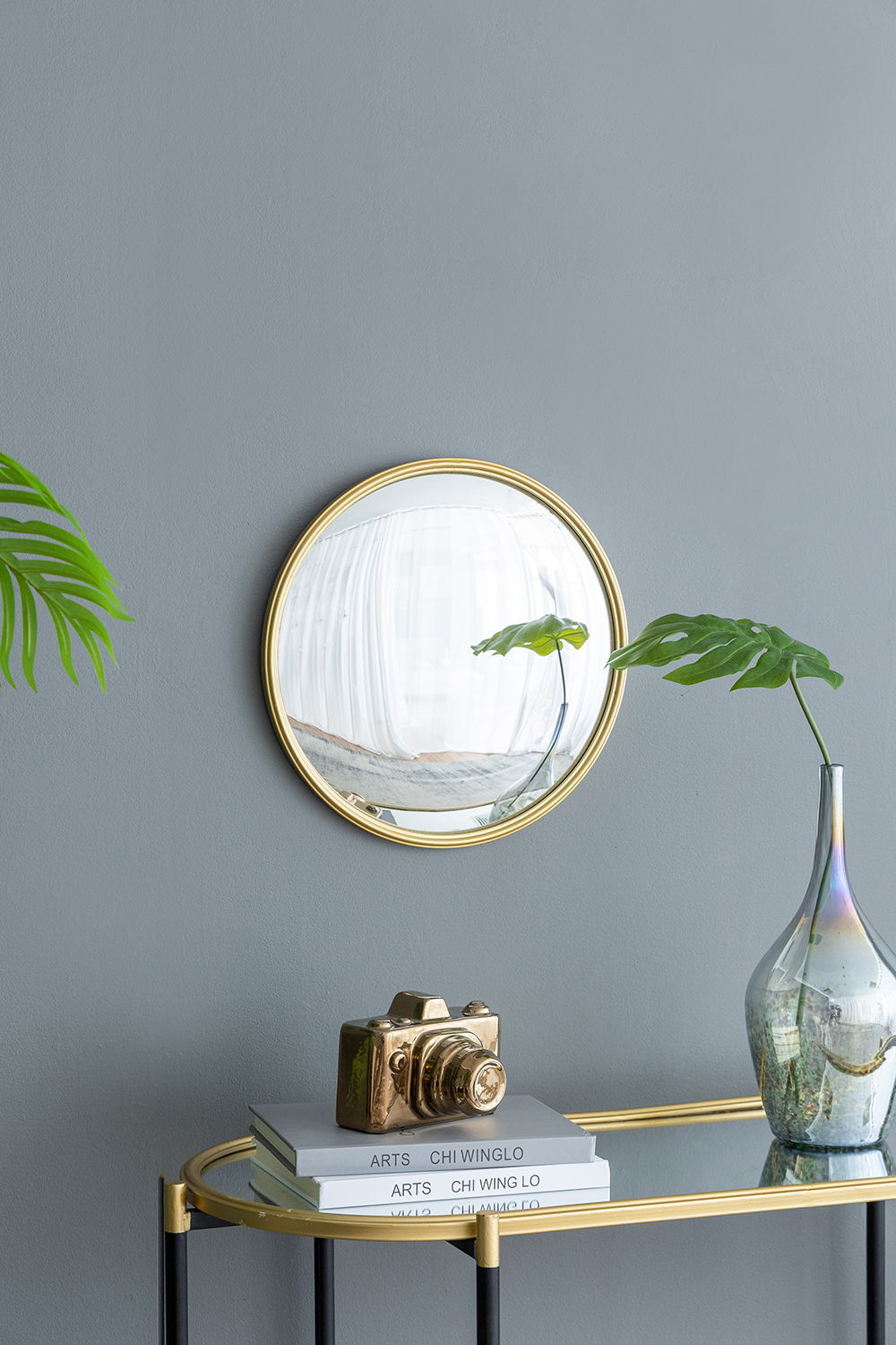 A Round Mirror, Circle Mirror With Iron Frame For Living Room Bedroom Vanity Entryway Hallway hangs on a gray wall above a glass console table holding stacked books, a gold camera ornament, a glass vase with green leaves, and a partially visible potted plant.