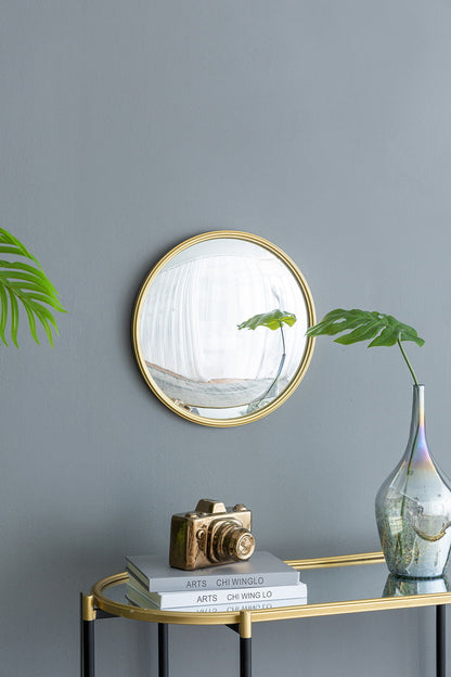 A Round Mirror, Circle Mirror With Iron Frame For Living Room Bedroom Vanity Entryway Hallway hangs on a gray wall above a glass console table holding stacked books, a gold camera ornament, a glass vase with green leaves, and a partially visible potted plant.