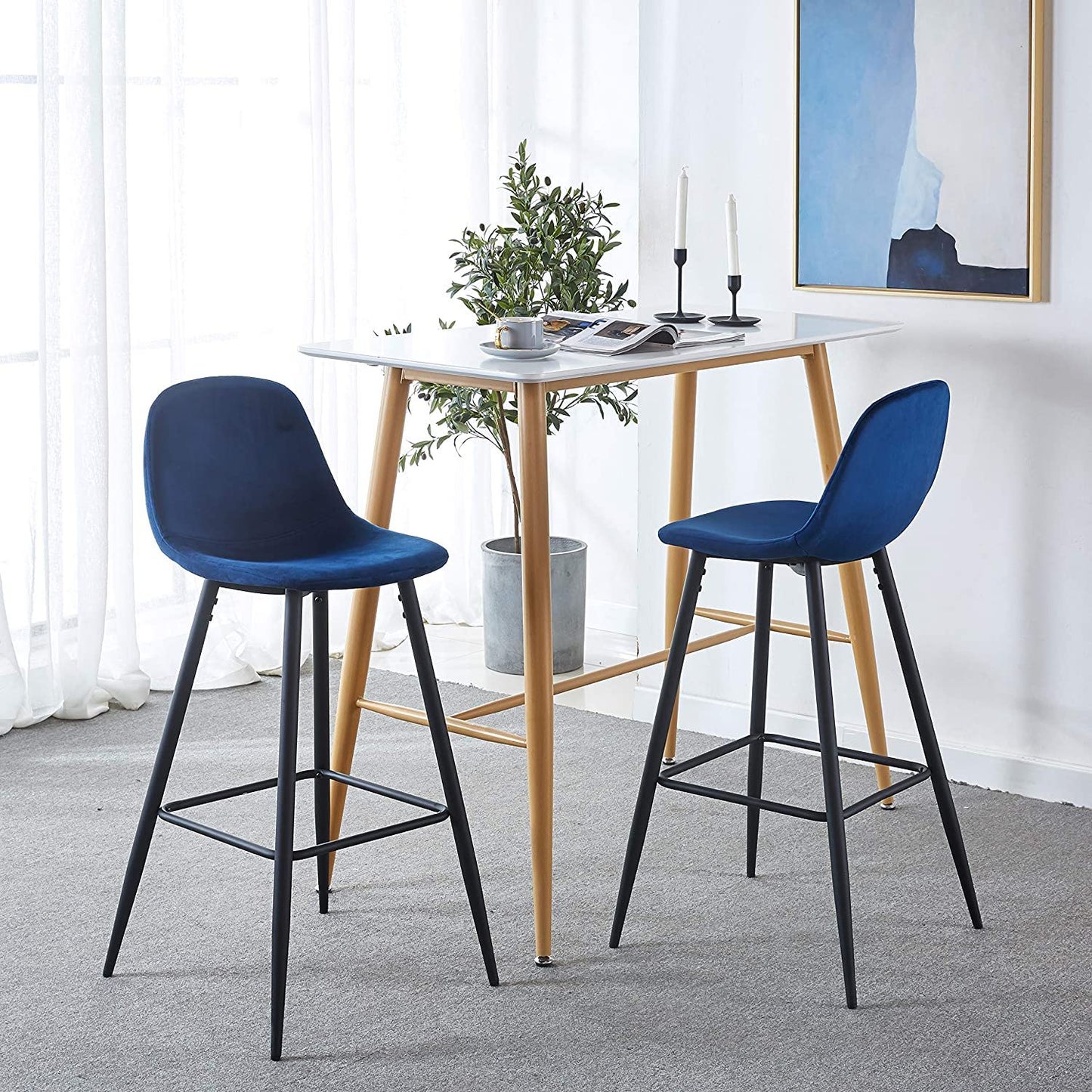 A modern dining space featuring a small square wooden table, two Velvet Barstool With Back (Set of 2) in blue with black metal legs, candles, greenery, and tableware in a light-filled room accented by framed abstract art.