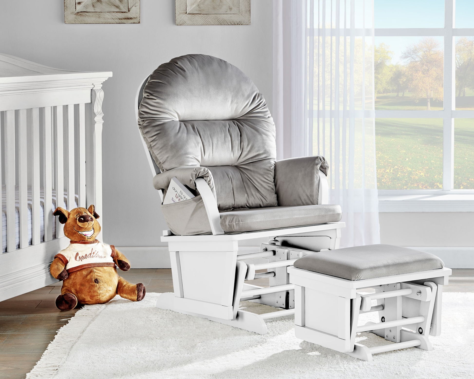 A cozy nursery includes the Madison Glider & Ottoman with a white base, a white crib, a plush brown dog toy in a "Goodies" shirt, and sunlight streaming through sheer curtains on a large window.