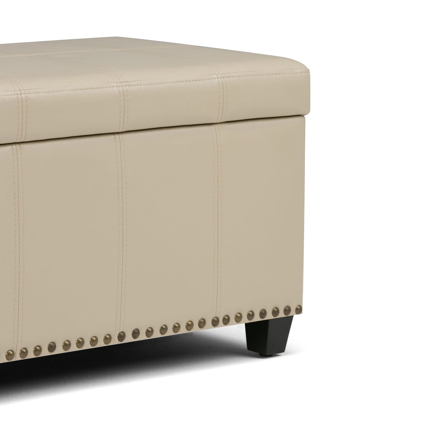 Close-up of the Amelia - Transitional Storage Ottoman Bench featuring beige upholstery, visible stitching, black legs, and decorative metal studs along the bottom edge against a white background.