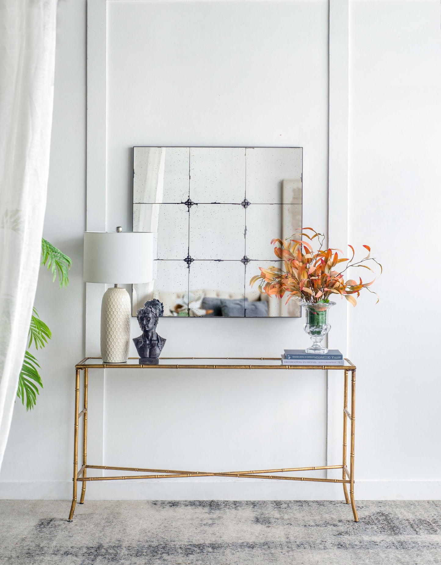 A gold console table with a white lamp, black bust, and orange flowers sits below the Traditional Decor Style Antique Glass Panel Mirror on a white wall; a leafy plant and sheer curtain are visible on the left.