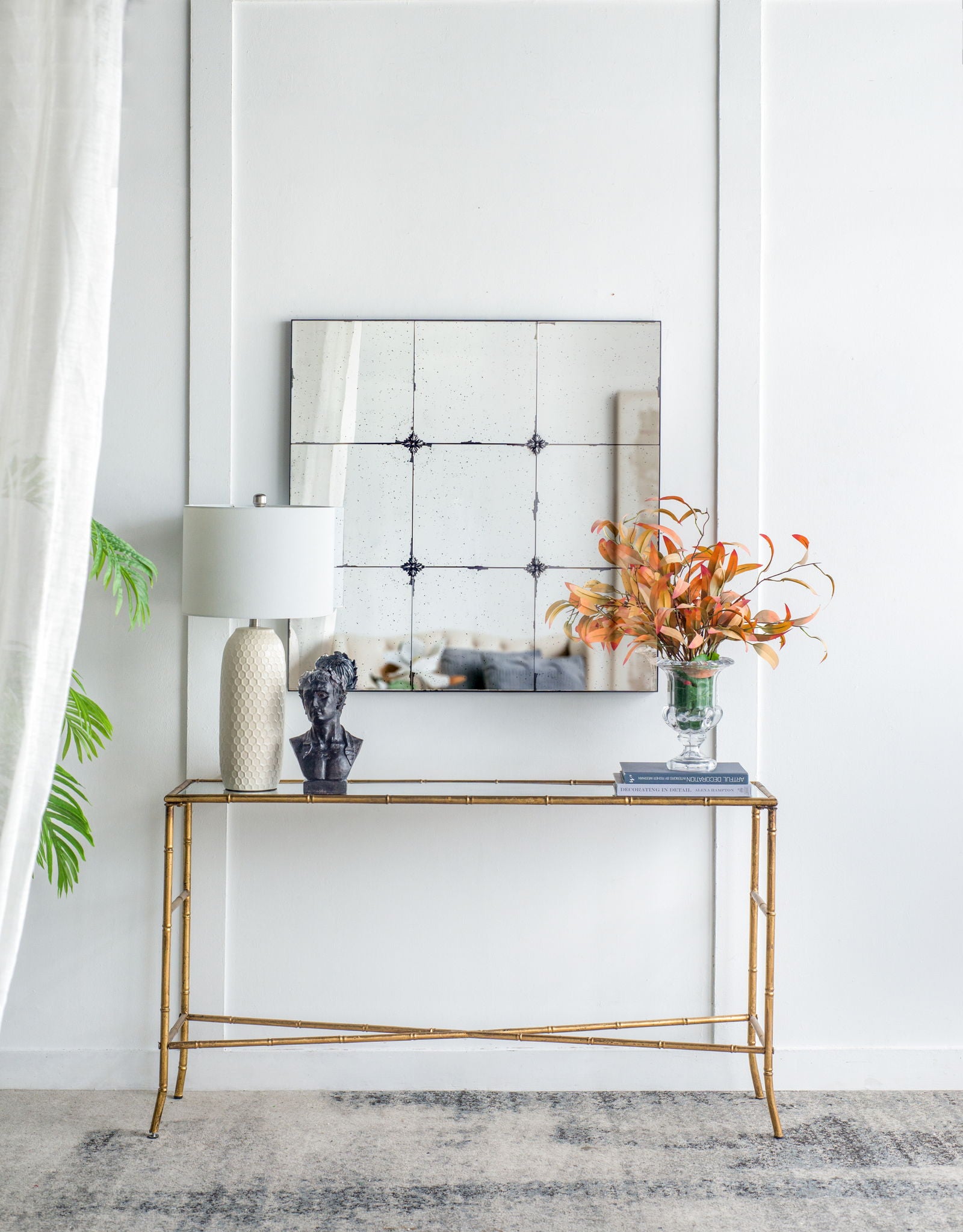 A gold console table with a white lamp, black bust, and orange flowers sits below the Traditional Decor Style Antique Glass Panel Mirror on a white wall; a leafy plant and sheer curtain are visible on the left.
