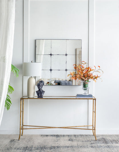 A gold console table with a white lamp, black bust, and orange flowers sits below the Traditional Decor Style Antique Glass Panel Mirror on a white wall; a leafy plant and sheer curtain are visible on the left.