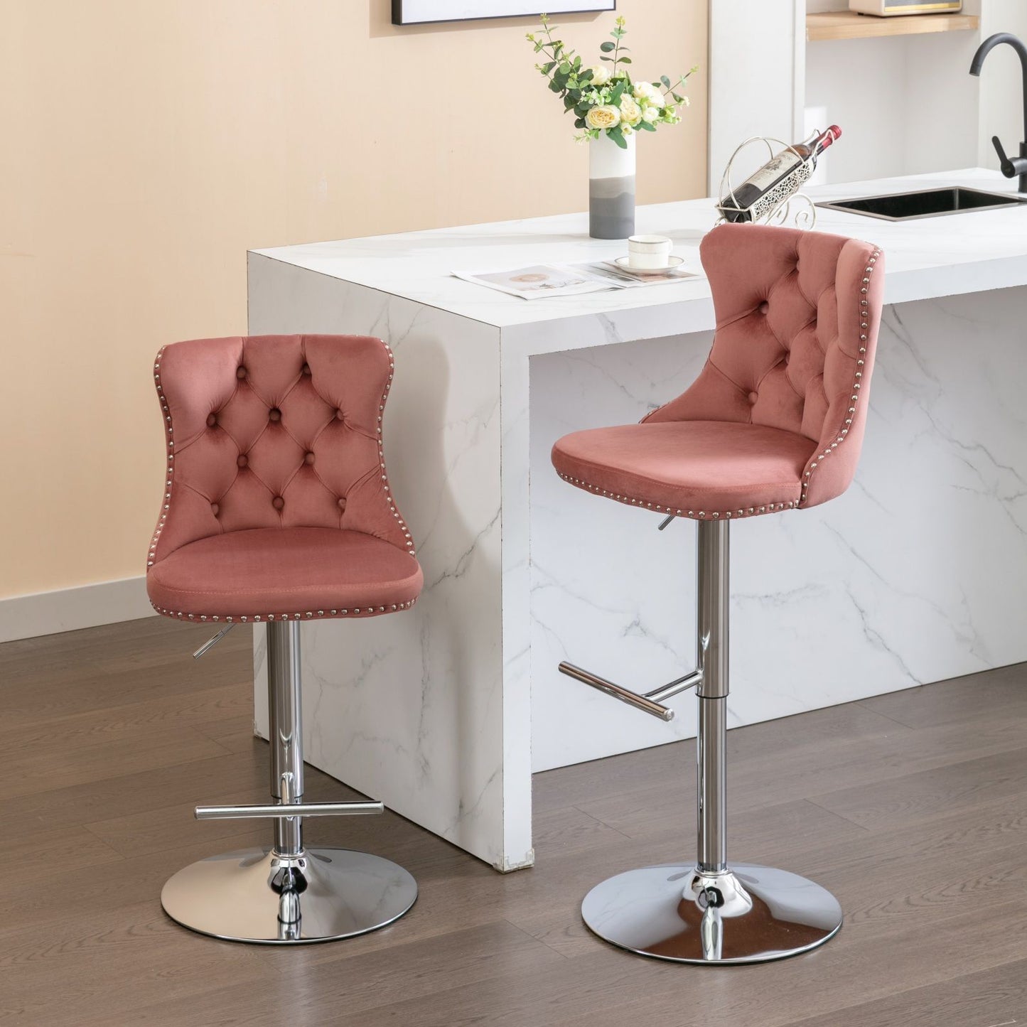 A set of two Swivel Velvet Barstools with tufted backs, chrome bases, and adjustable seat height (25-33") are placed by a white marble kitchen island featuring a vase, wine holder, and a sink.