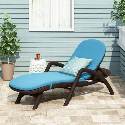 The Mikael - Chaise Lounge With Cushion, featuring a blue cushion and striped pillow, sits on a patio near a white side table with a drink. Two potted plants are by a light blue house with a window in the background.
