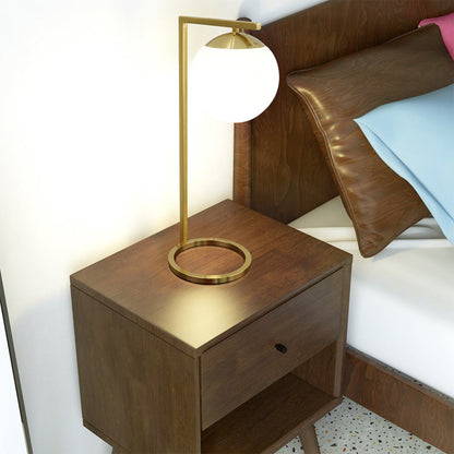 A modern wooden nightstand with a drawer and open shelf holds the Globe Table Lamp Opal With Dimmer Switch Inline in Gold/White, beside a bed with brown pillows and light blue and pink bedding.