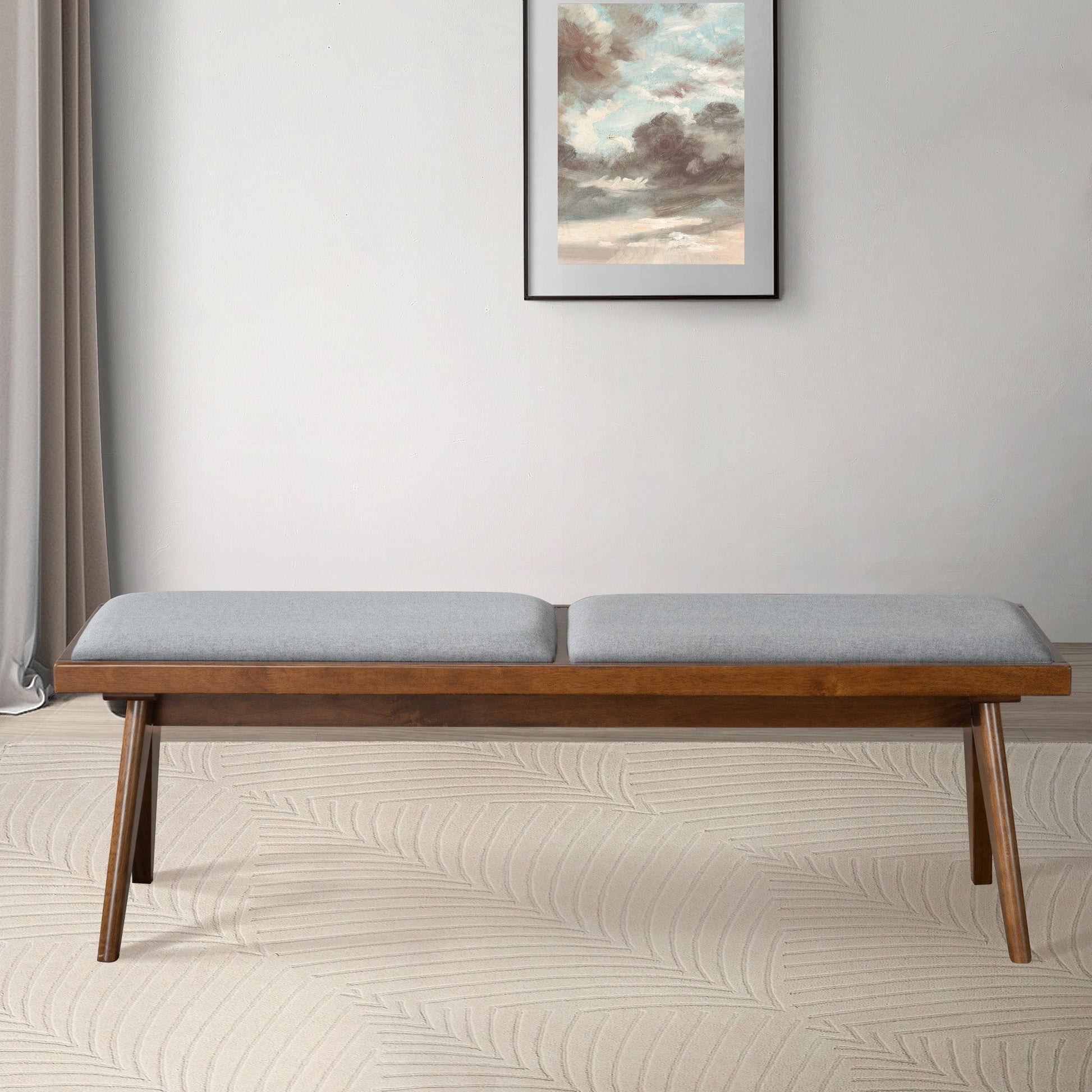 The Keira - Modern Aesthetics Bench with a light gray cushion sits on a beige patterned rug before a light wall accented by a framed cloud painting, with sheer curtains draped to the left.