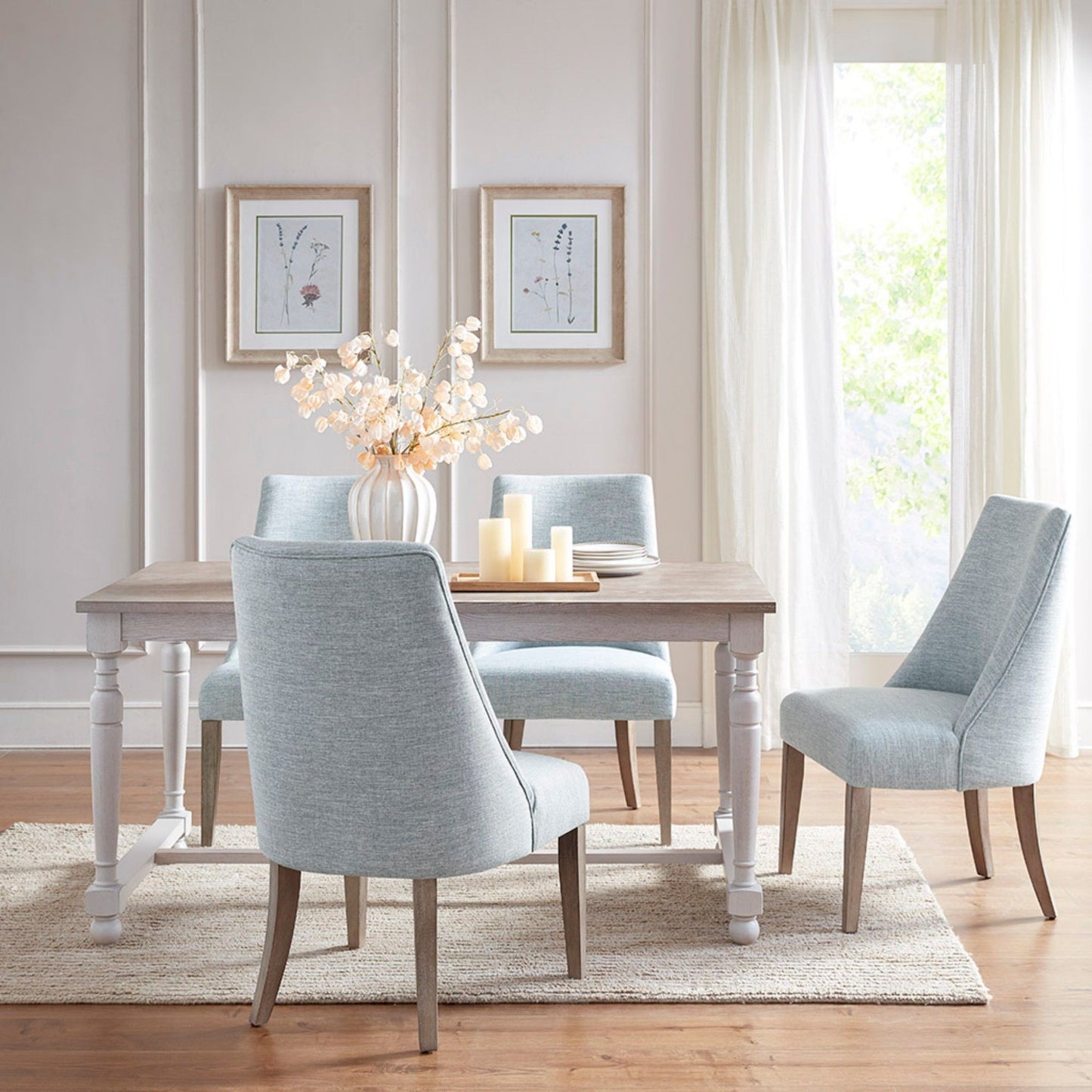 A bright dining room with a light wood table, two sets of Comfortable Dining Chair Upholstered in blue, a vase of white flowers, three candles, and framed botanical prints on pale walls. Sheer curtains frame a sunlit window.