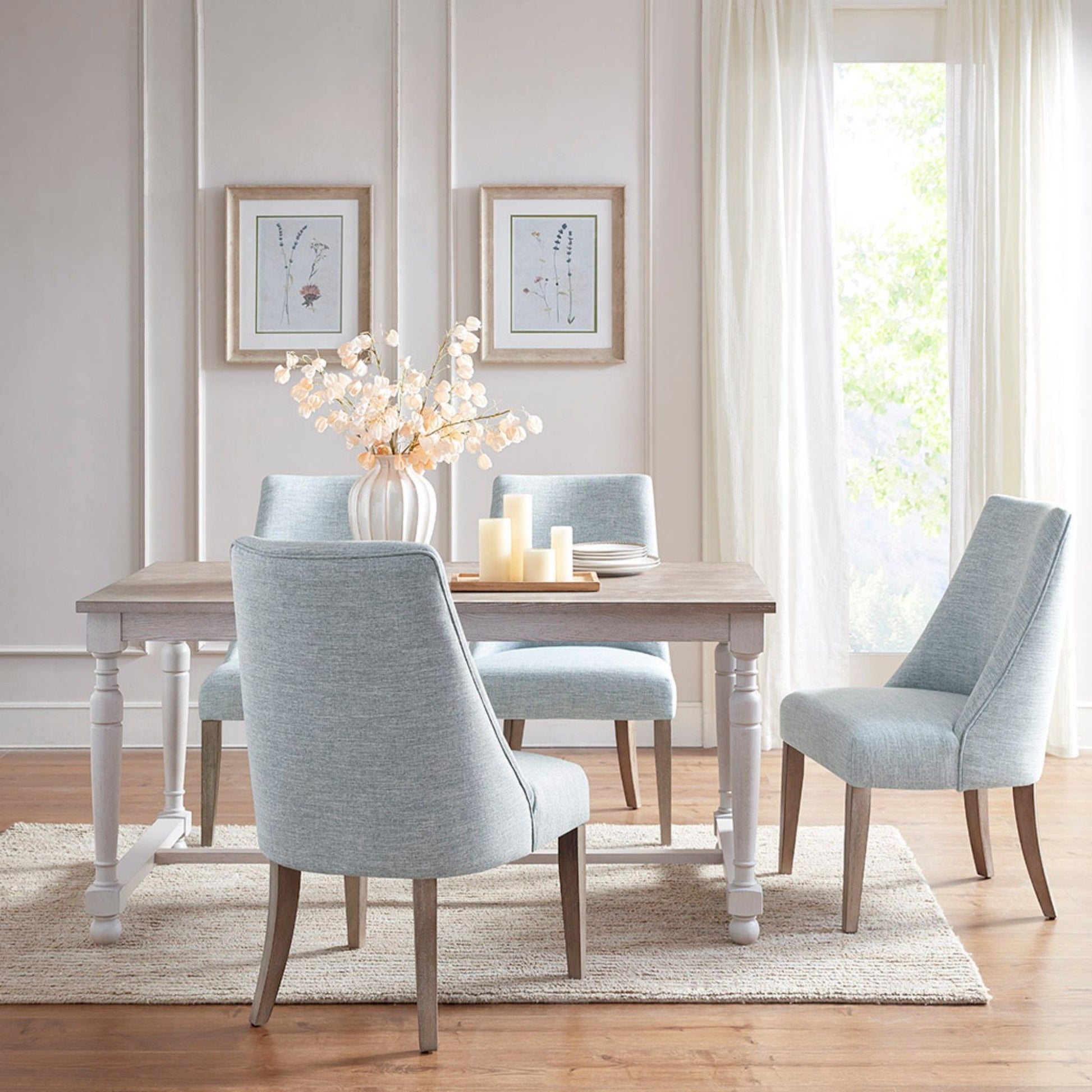 A bright dining room with a light wood table, two sets of Comfortable Dining Chair Upholstered in blue, a vase of white flowers, three candles, and framed botanical prints on pale walls. Sheer curtains frame a sunlit window.