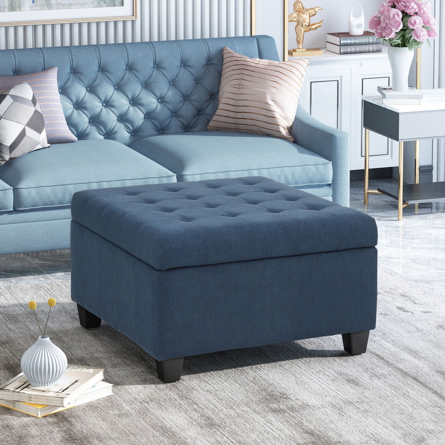 The Isabella Contemporary Ottoman With Storage in blue sits before a matching sofa with decorative pillows in a stylish living room, complete with light décor, a side table, books, and a vase on a rug.