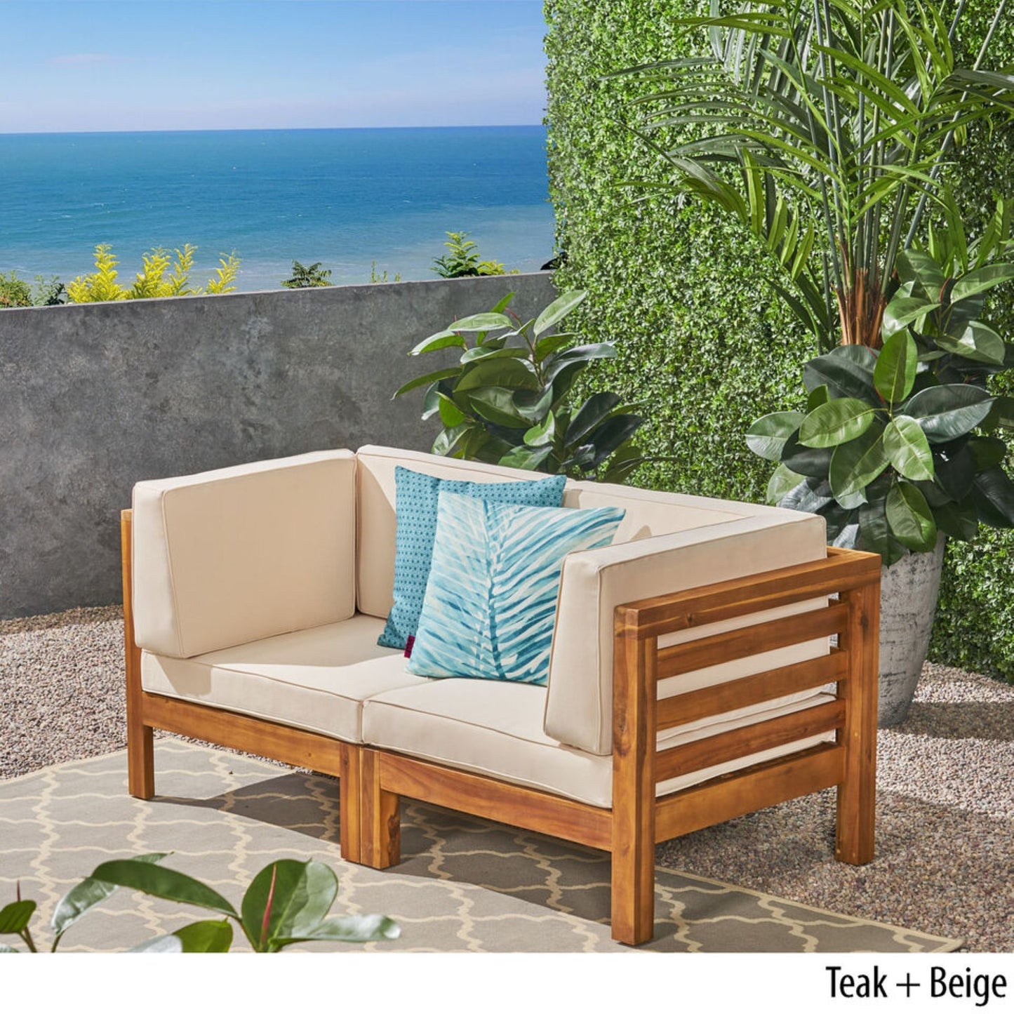 The Oana 2 Piece Loveseat Set with Water Resistant Cushions in teak wood and beige sits on a patio, accented by potted plants, a green hedge wall, ocean views, and two blue decorative pillows.
