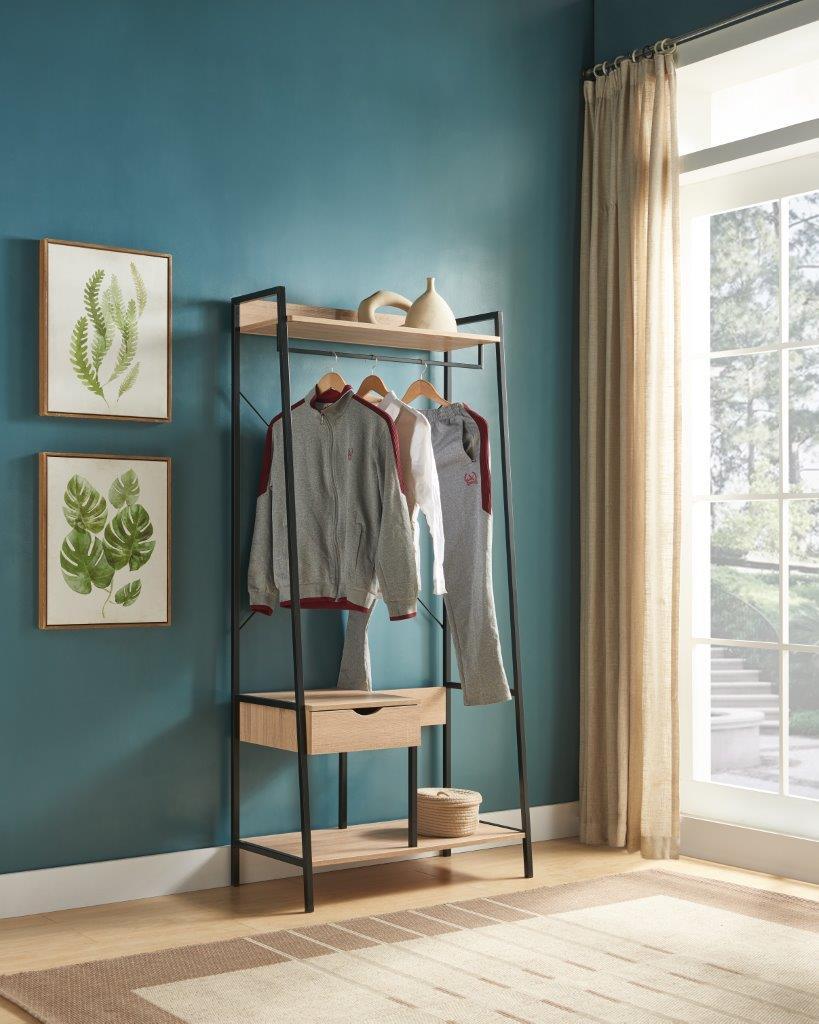 The Metal Clothes Rack Open Wardrobe Free Standing With One Drawer features a hanging rod displaying a shirt and pants on wooden hangers. Positioned by a teal wall, it’s accented by leaf print art, beige curtains, and sunlight from a large window.