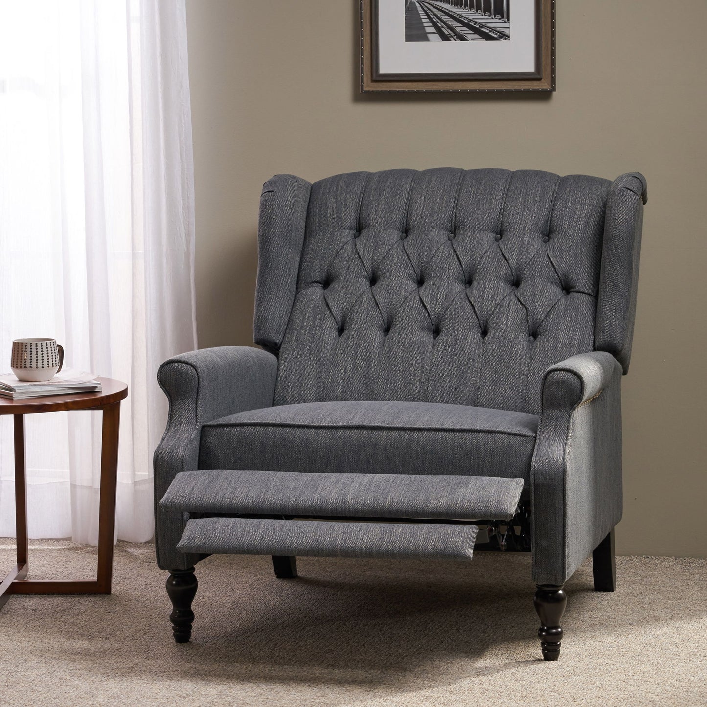 A Dunham Contemporary Fabric Tufted Oversized Pushback Recliner in gray sits on a carpeted floor beside a round wooden table with a mug, near a window with sheer curtains and a framed photo on the wall.
