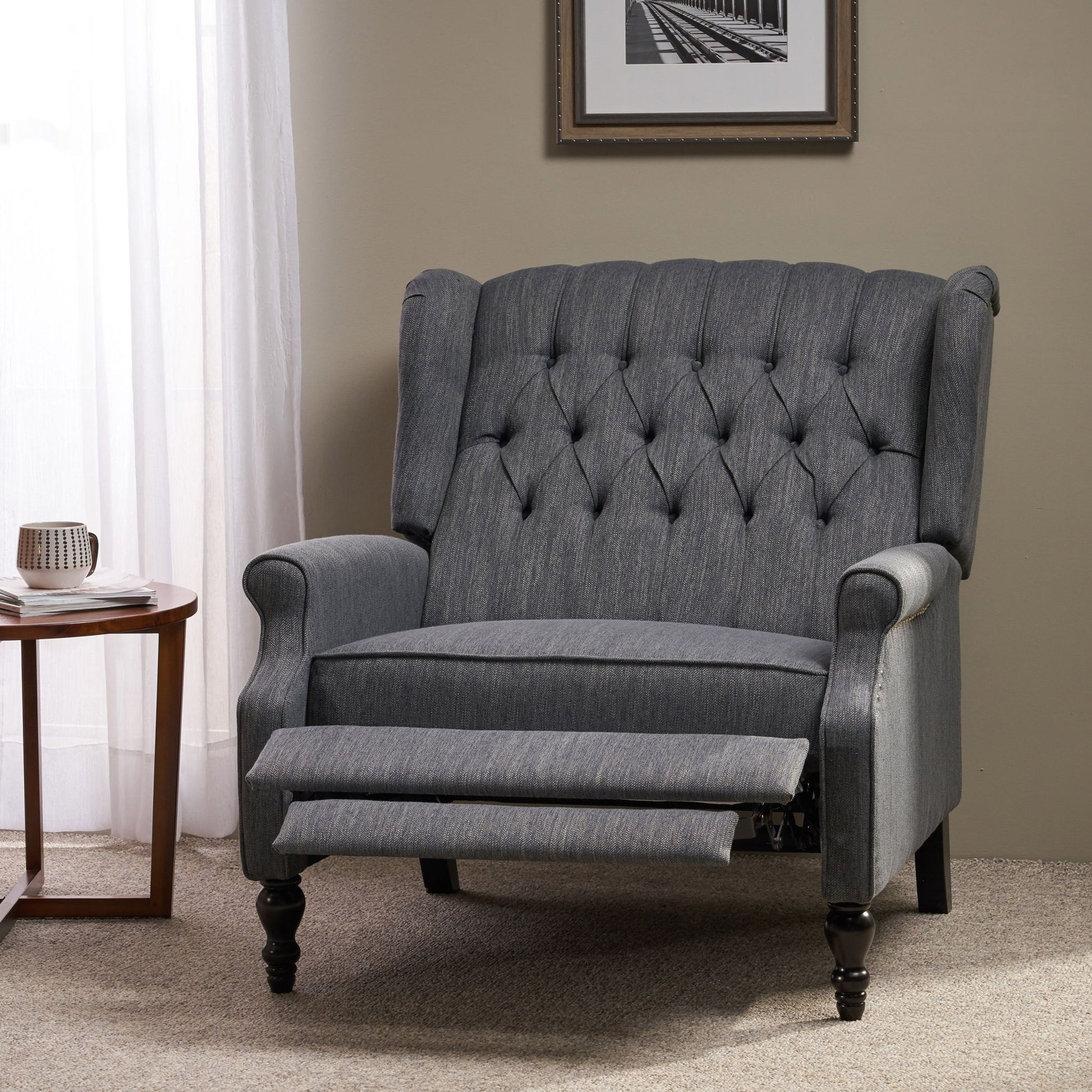 A Dunham Contemporary Fabric Tufted Oversized Pushback Recliner in gray sits on a carpeted floor beside a round wooden table with a mug, near a window with sheer curtains and a framed photo on the wall.
