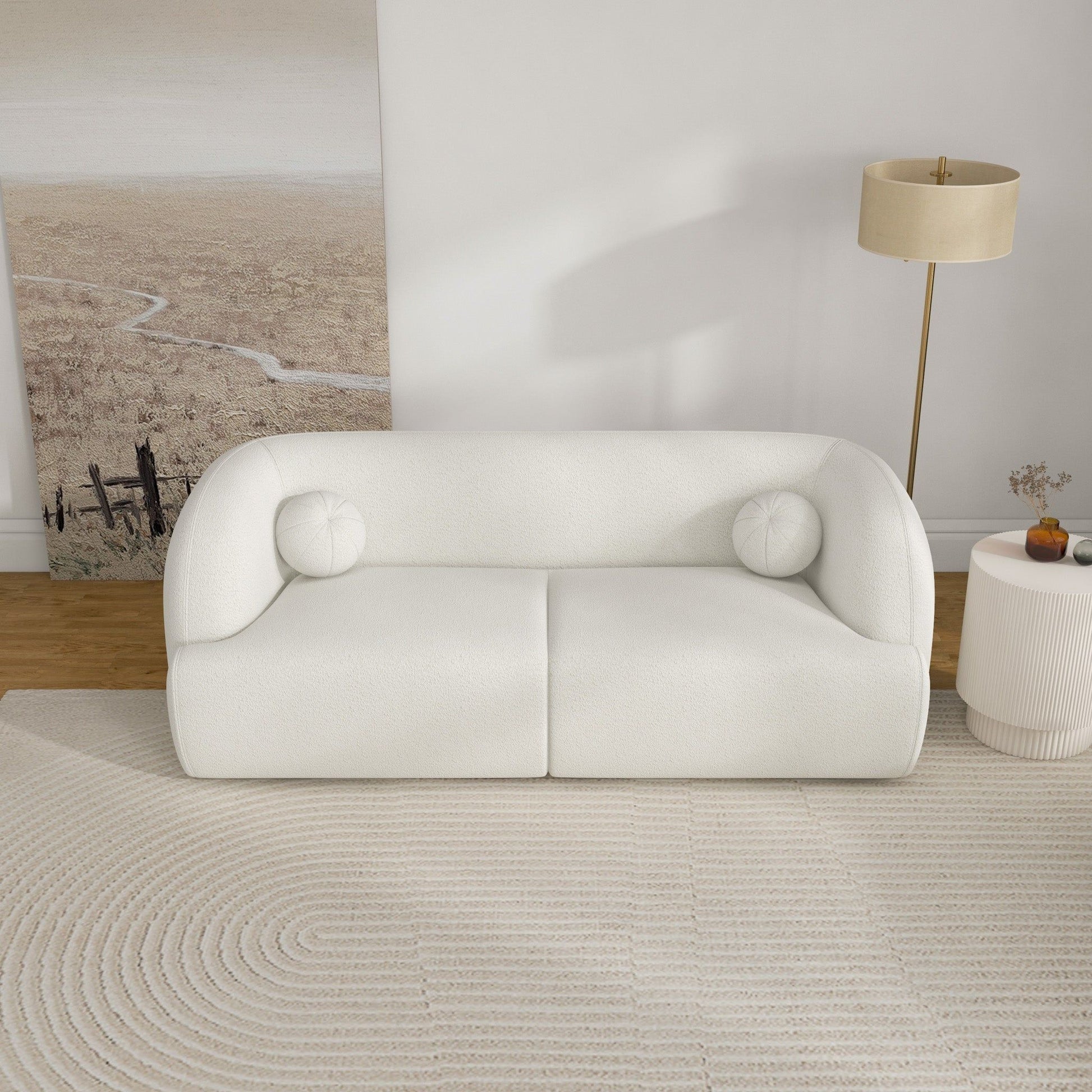 A modern living room featuring the Anna - French Boucle Sofa, two round cushions, a textured beige rug, a round side table with decor, a tall floor lamp, and a large landscape art piece on the wall.