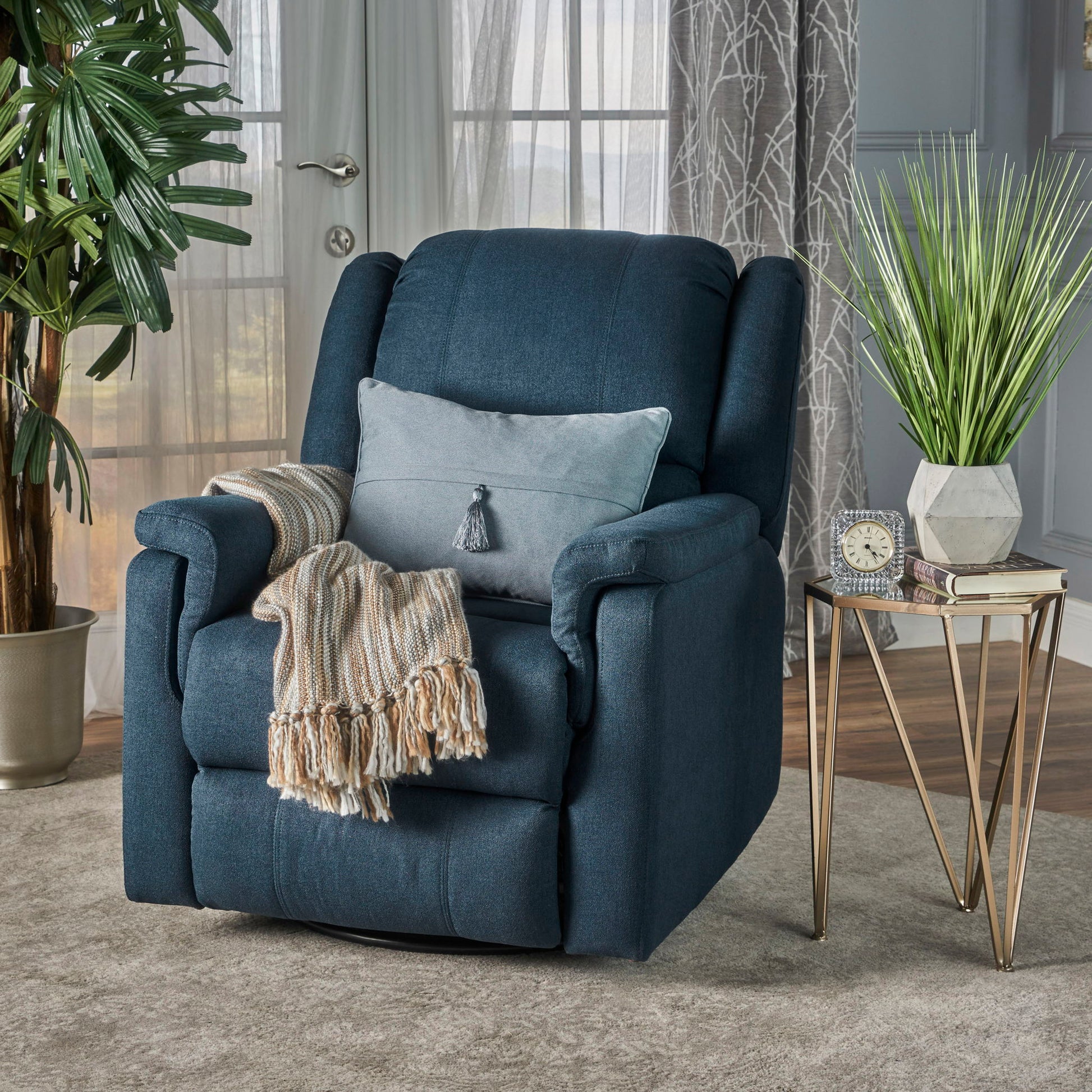A Contemporary Glider Recliner With Swivel Function in navy blue, topped with a light grey pillow and beige striped throw, sits on a light rug beside a gold and glass side table holding plants, books, and a clock in a cozy, sunlit living room.