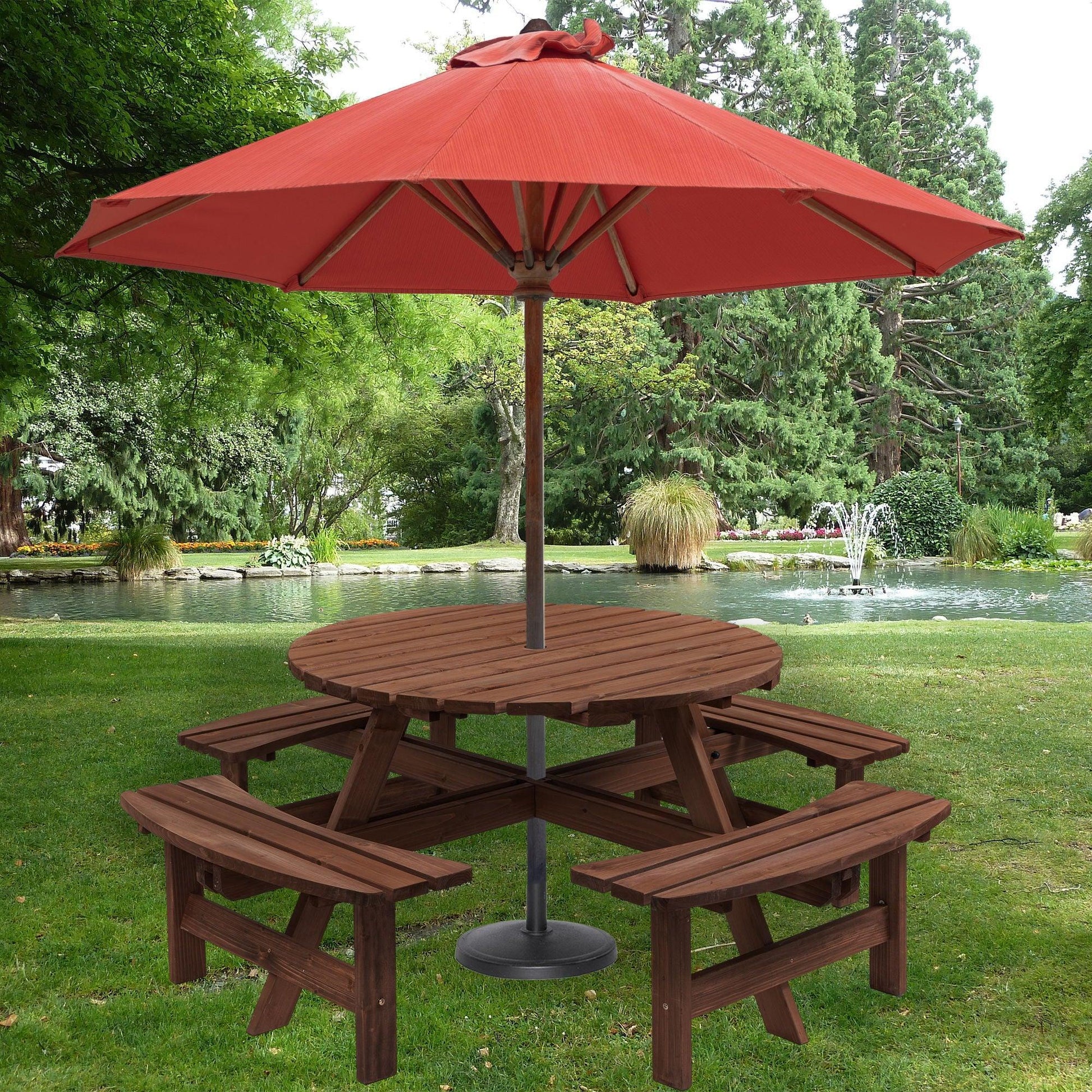 An 8 Person Wooden Picnic Table with 4 built-in benches sits on green grass under a large red umbrella, with a pond, fountain, and lush trees in the background.