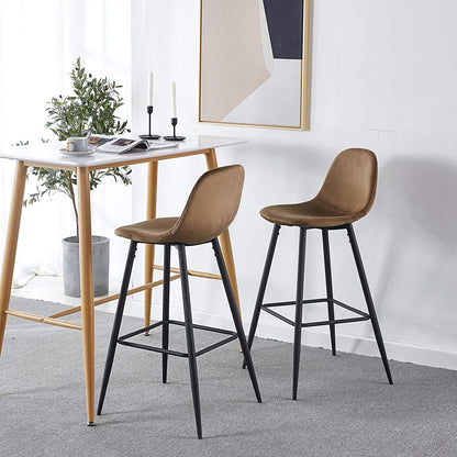 Two Velvet Barstool With Back (Set of 2) in brown with black legs sit by a wooden table adorned with plates, cups, and candles. A plant and abstract art complete the bright, minimalist room.