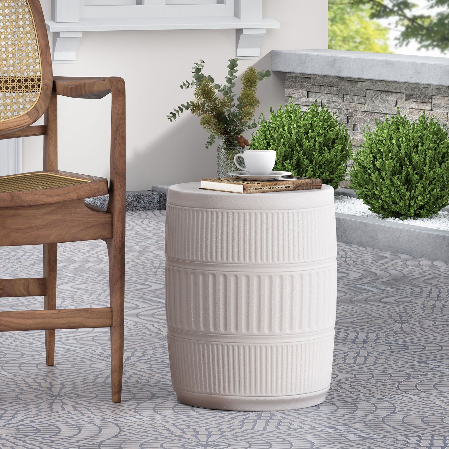 The Outdoor Patio End Table - Antique White, round and ceramic, holds a cup, saucer, and book on a patterned patio beside a wooden chair, with green shrubs and a stone border in the background.