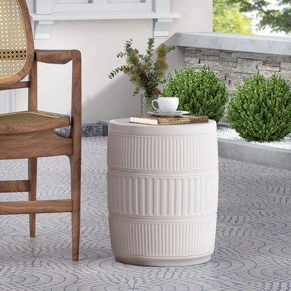 The Outdoor Patio End Table - Antique White, round and ceramic, holds a cup, saucer, and book on a patterned patio beside a wooden chair, with green shrubs and a stone border in the background.