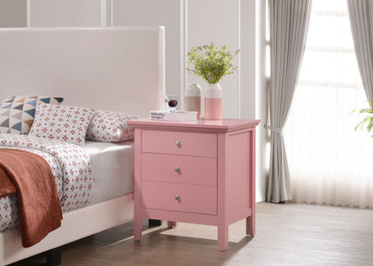 The Hammond 3 Drawer Nightstand in pink stands beside a bed with patterned bedding. Two vases with green plants and a wine glass sit on the nightstand. Sunlight streams through a window with gray curtains.