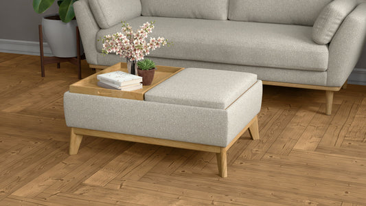 The Revel Reversible Tray Ottoman in tan sits on a wooden herringbone floor, holding a tray with a pink flower arrangement, small plant, and closed book. A matching sofa and greenery appear in the background.