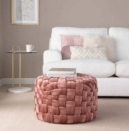 A cozy living room featuring a white sofa, pink and white pillows, a round glass side table with a white mug, and the Braided - Versatile Round Ottoman in pink, topped with books in the foreground.