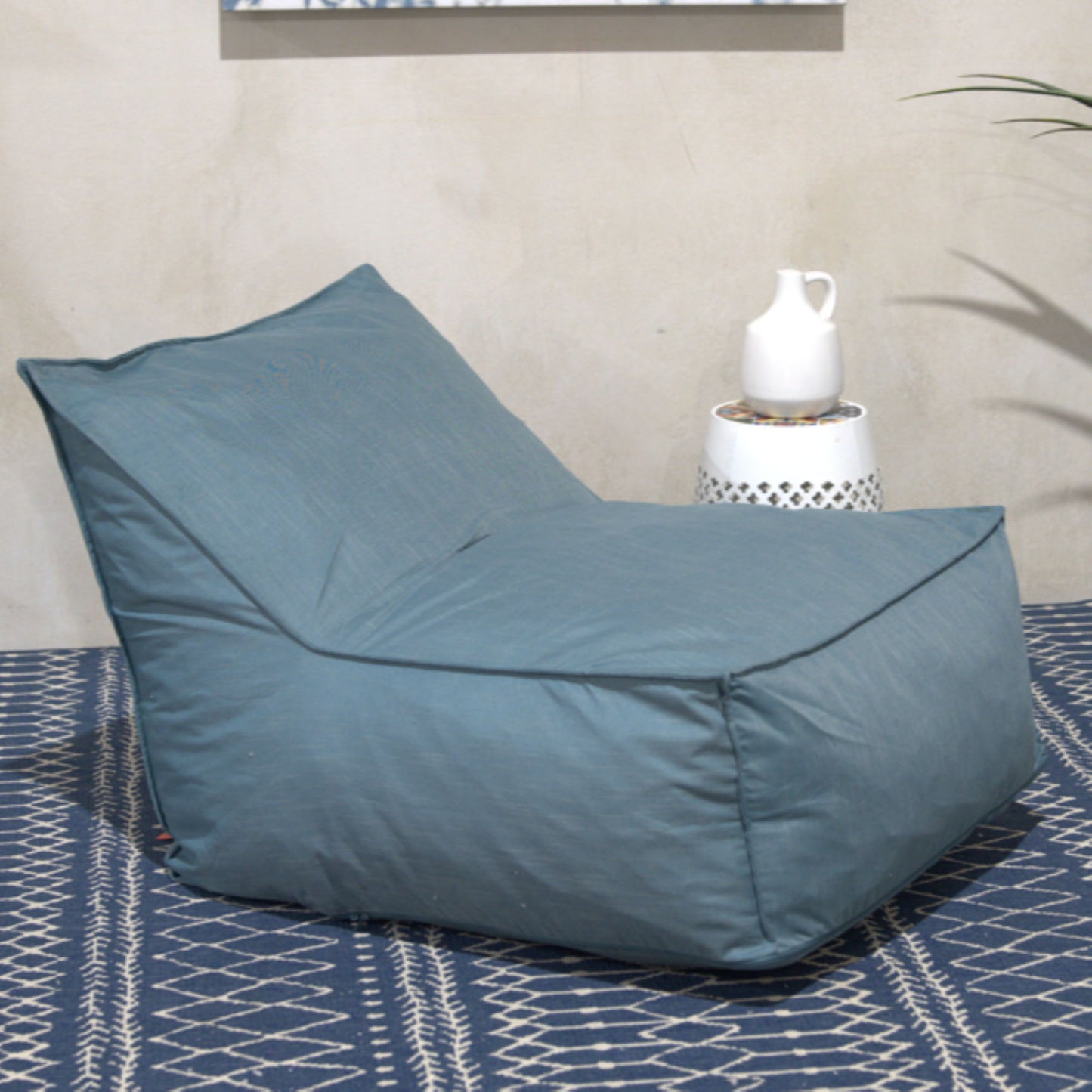 The Ettie Water Resistant Fabric Bean Bag Chair in blue sits on a patterned blue rug next to a white side table with a white ceramic pitcher, all set against a light-colored wall.