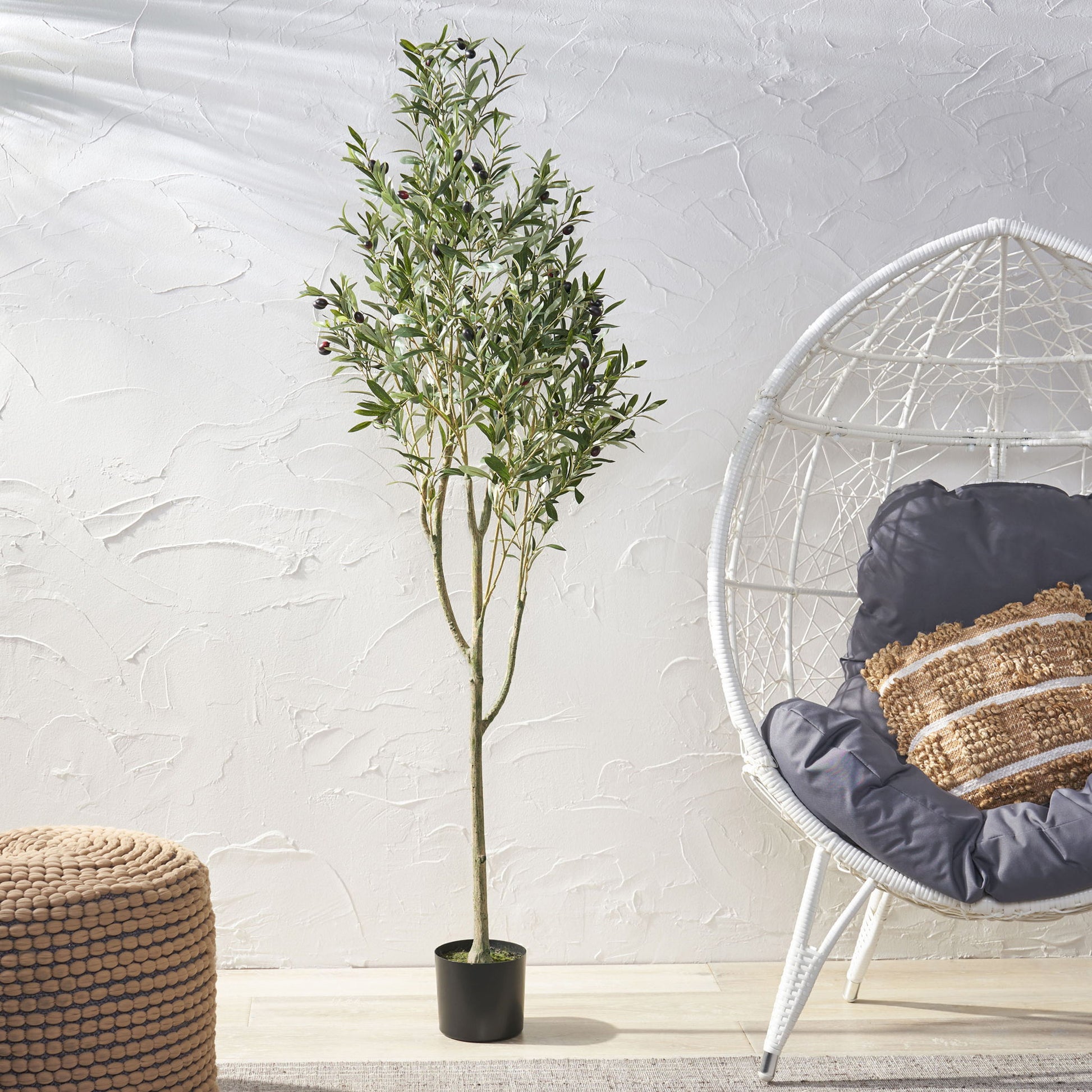 An Artificial Olive Tree stands indoors next to a woven pouf and a white egg-shaped chair with dark cushions and a decorative pillow, all set against a textured white wall.