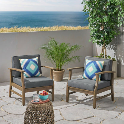 Two Perla Contemporary Club Chairs with water resistant cushions and blue patterned pillows sit on a stone terrace overlooking the ocean, beside a small table with fruit and a drink, and nearby potted plants.