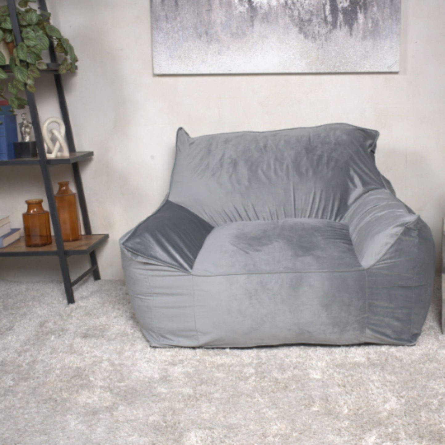 The Allea - Bean Bag Chair With Armrests, plush and gray, sits on a soft gray carpet in a modern living room. Nearby are shelves with decor, a leafy plant, and abstract art hanging above.