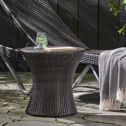 The Thrive Hourglass Rattan Table, without a glass top, holds a glass of water with lime and a closed book. A matching lounge chair and fringed blanket sit nearby on a stone patio surrounded by greenery.
