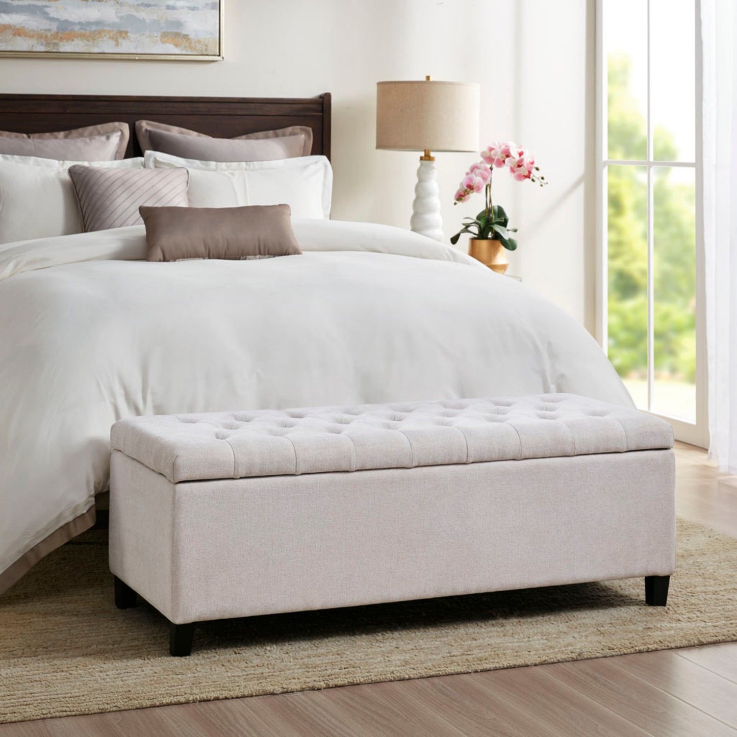 A neatly made bed with white bedding and a brown headboard sits in a bright bedroom. In front is an Elegant Design Tufted Top, Soft Close Storage Bench in light beige. A lamp, orchid, and sunlit window complete the scene.