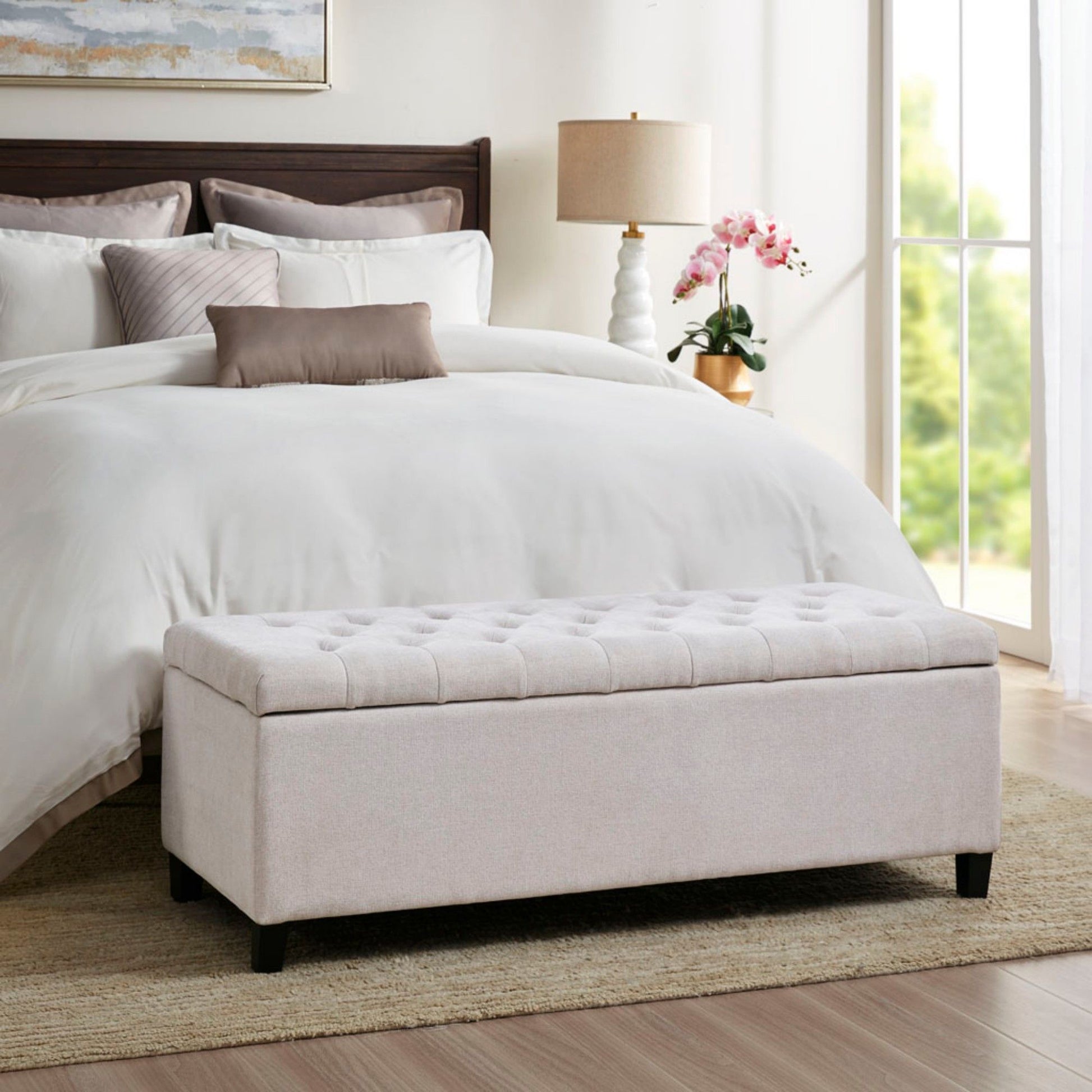 A neatly made bed with white bedding and a brown headboard sits in a bright bedroom. In front is an Elegant Design Tufted Top, Soft Close Storage Bench in light beige. A lamp, orchid, and sunlit window complete the scene.