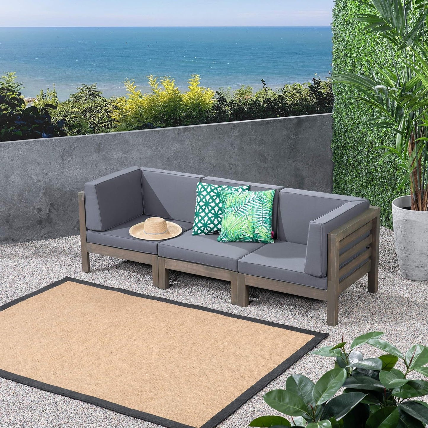 The Oana - 3 Piece Sofa Set With Water Resistant Cushions, featuring two green leaf-patterned pillows and a straw hat, sits on a gravel patio with ocean views, surrounded by plants and a beige rug.