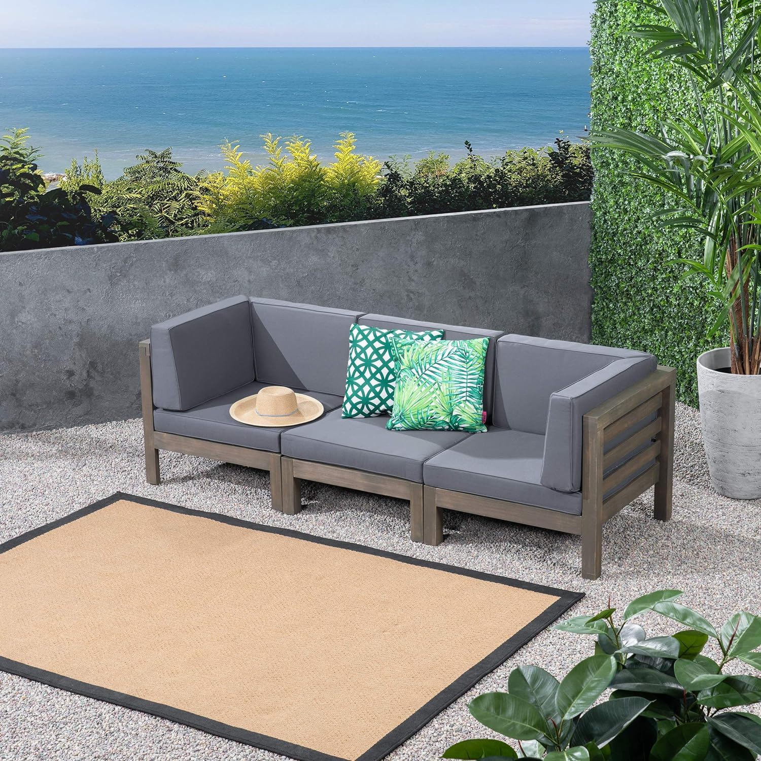 The Oana - 3 Piece Sofa Set With Water Resistant Cushions, featuring two green leaf-patterned pillows and a straw hat, sits on a gravel patio with ocean views, surrounded by plants and a beige rug.