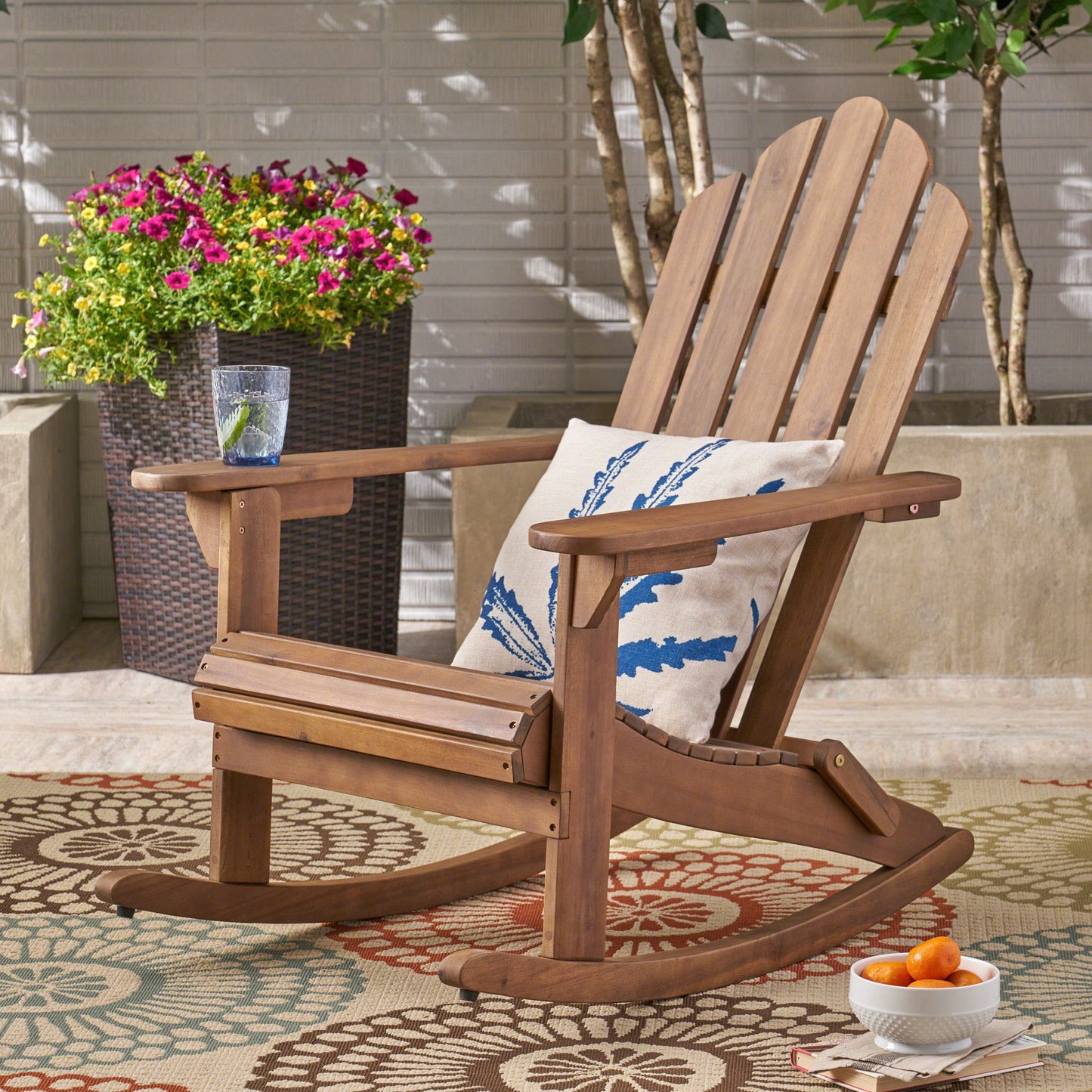 The Echoes Outdoor Acacia Wood Adirondack Rocking Chair with a leaf-print cushion rests on a patterned rug. A glass of water, apricots in a bowl, and potted flowers complete the bright patio scene.
