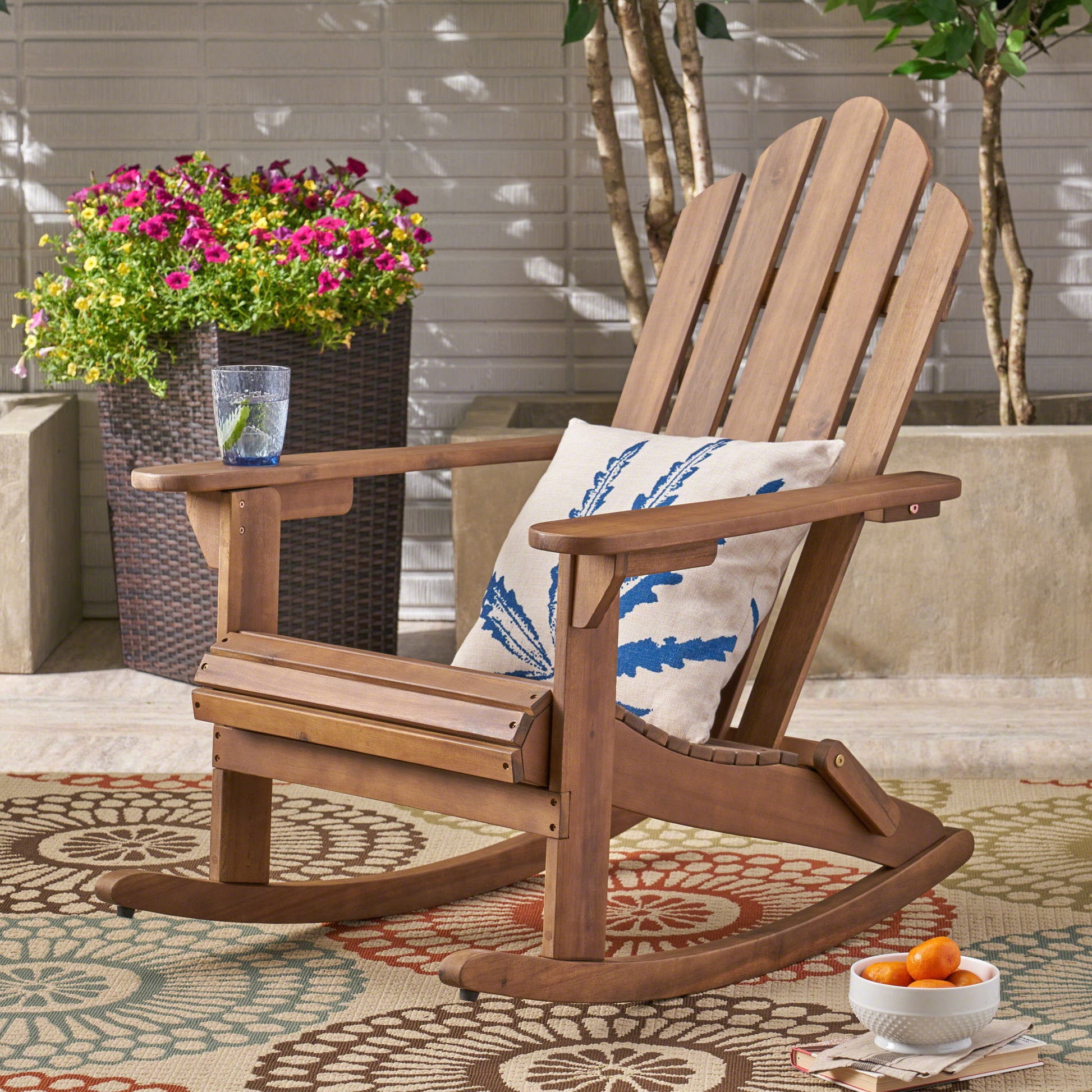 The Echoes Outdoor Acacia Wood Adirondack Rocking Chair with a leaf-print cushion rests on a patterned rug. A glass of water, apricots in a bowl, and potted flowers complete the bright patio scene.