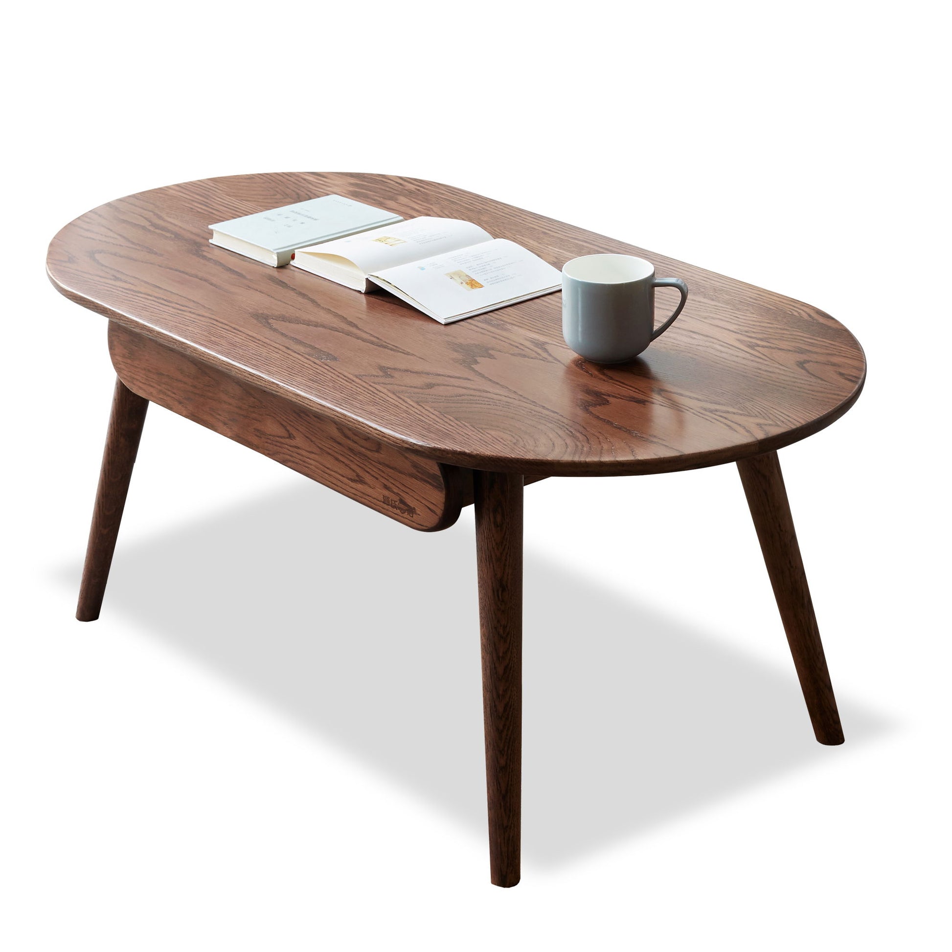 The Capsule Centre Low Table With Drawers Coffee Table, set against a white background, displays an open book, a closed book, and a gray mug neatly arranged on its polished oval wooden surface with angled legs.
