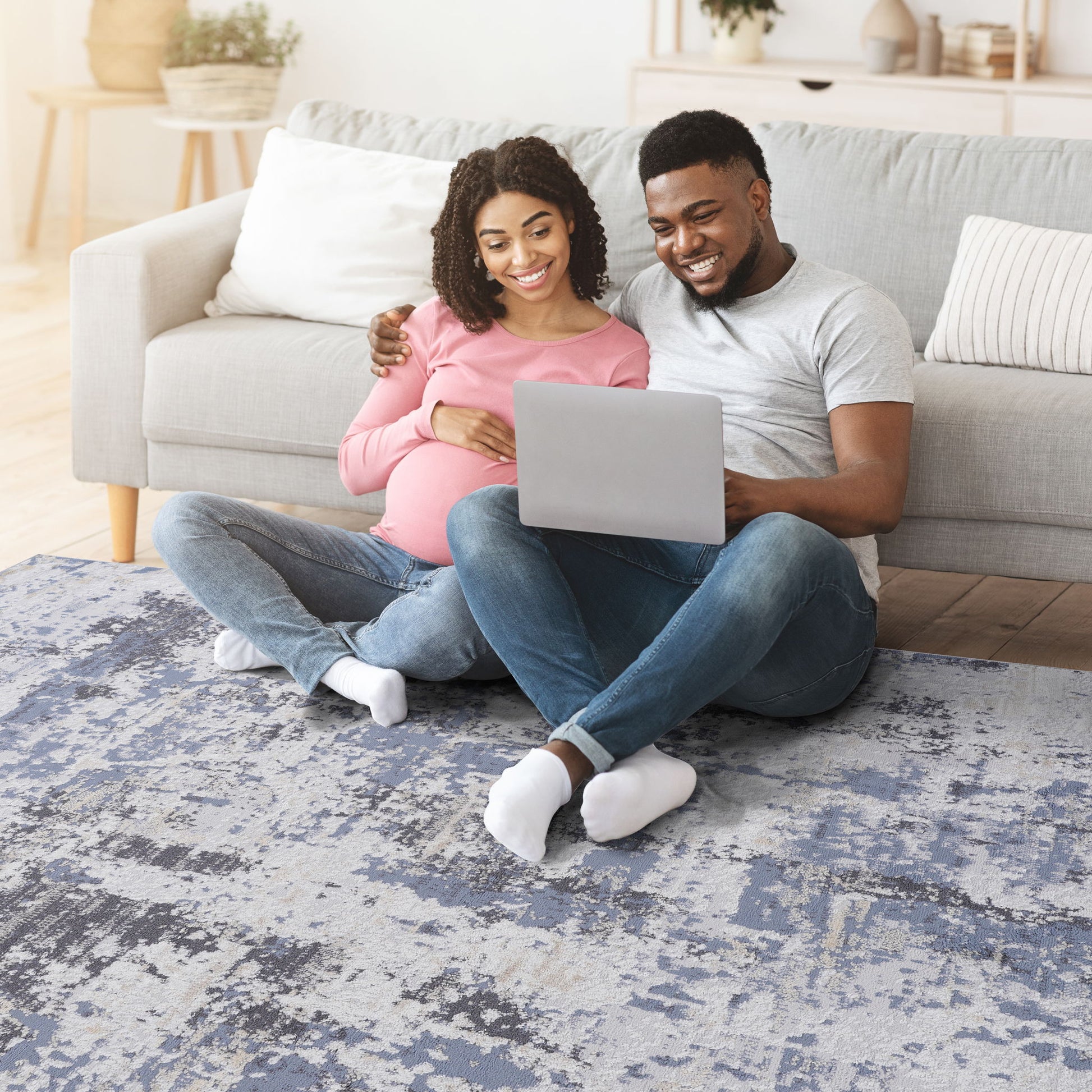 A pregnant woman and a man sit on the cozy Payas - Abstract Area Rug, leaning against a couch and smiling at a laptop, looking happy and relaxed in their living room.