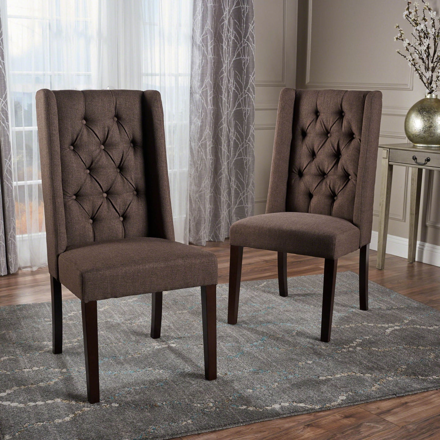 Two Sophisticated Tufted Dining Chairs With High Back Design in brown upholstery are arranged on a gray patterned rug in a modern room featuring wood flooring, light walls, sheer curtains, and a decorative table with a vase of flowers.