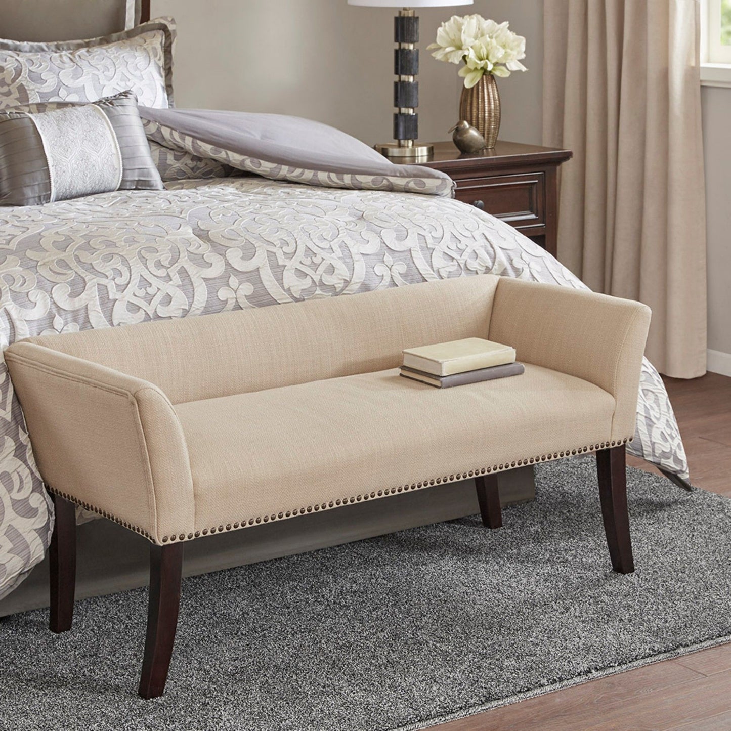 An Elegant Design Bench Upholstery with a solid wood frame sits at the foot of an ornate bed with gray and white bedding. A closed book rests on the bench, while a nightstand, lamp, and flowers are beside the bed.