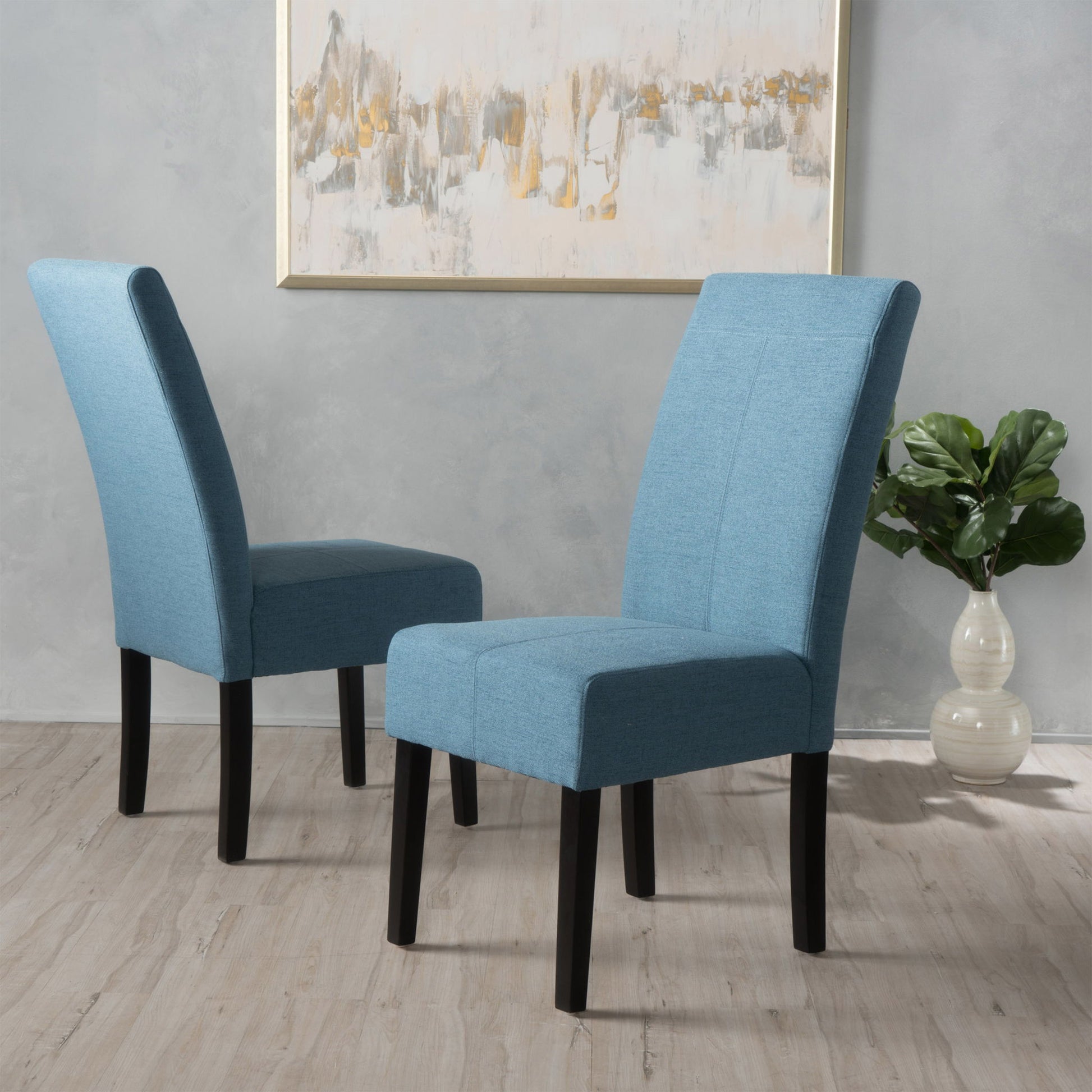 Two Pertica Dining Upholstered Armless Chairs (set of 2) with blue fabric and black legs sit on a light wood floor. A green potted plant in a white vase and an abstract painting decorate the light gray wall in the background.