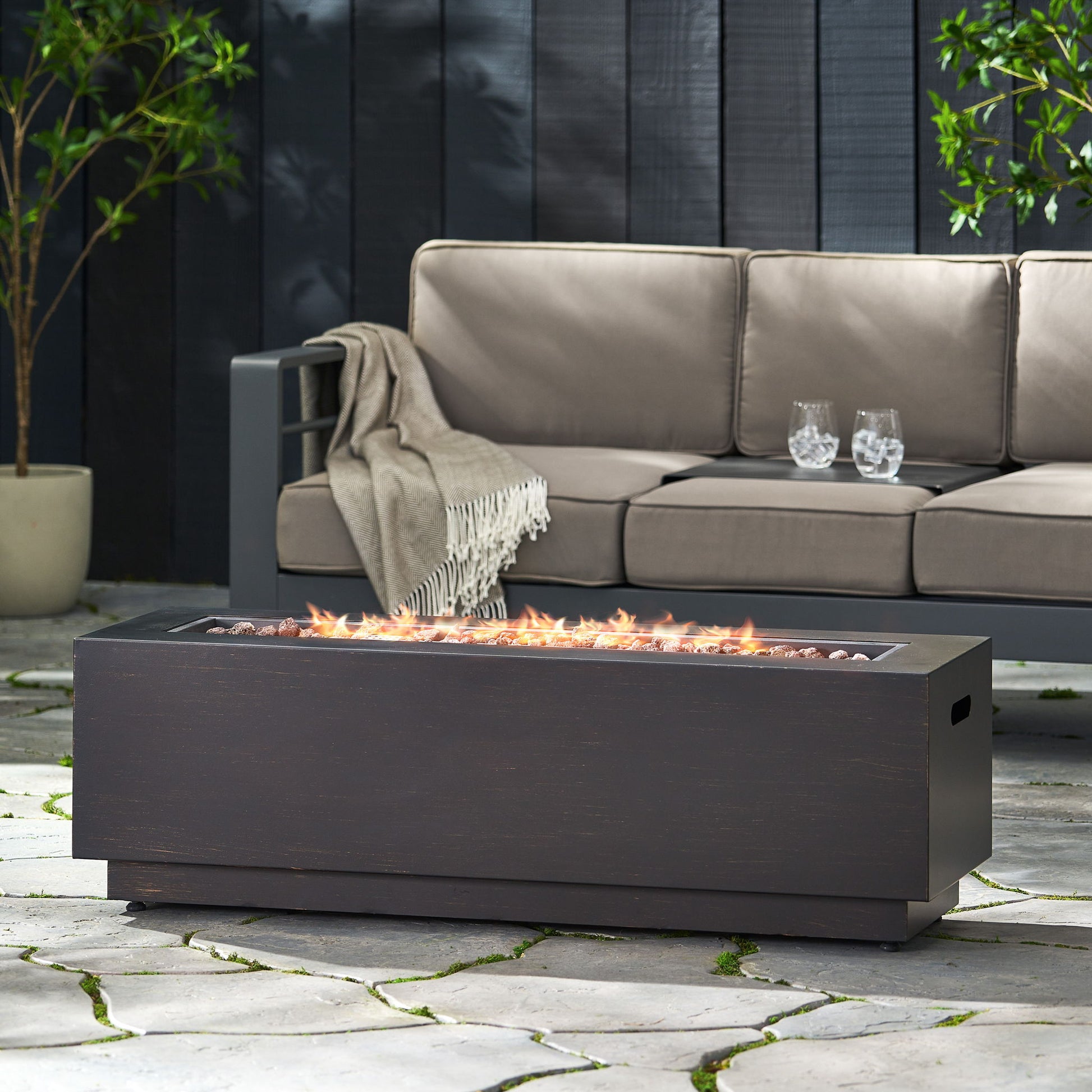A modern outdoor patio with a gray cushioned sofa, a beige throw, two glasses of water on the armrest, and the Modern Rectangular Iron Fire Pit 50,000 Btu burning bright on stone tiles dotted with green moss.
