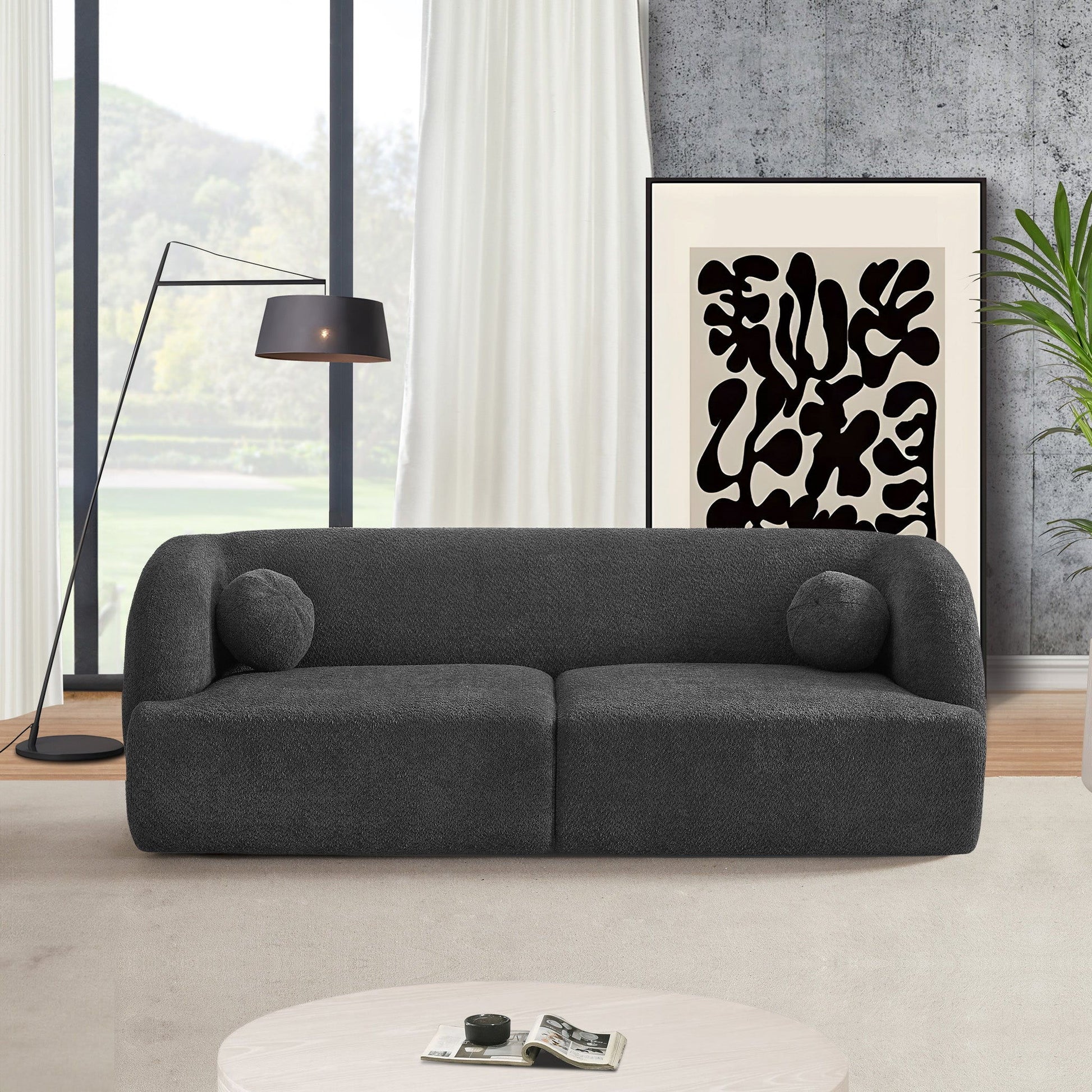 A modern living room with the Anna - French Boucle Sofa in dark gray, two round cushions, a black floor lamp, abstract black-and-white art, large window with white curtains, and a round white coffee table.